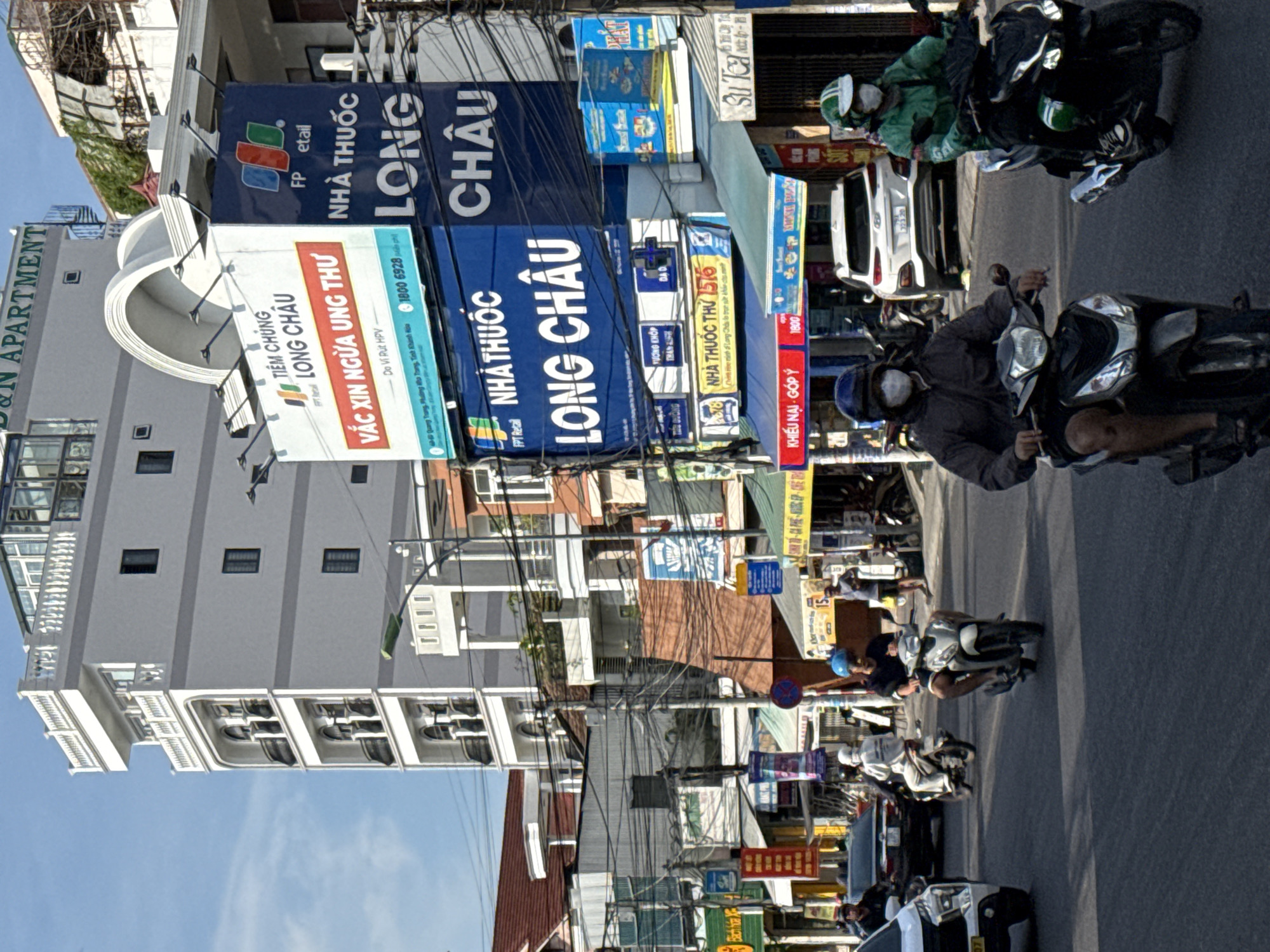 The image depicts a busy street scene in Bac Nha Trang Ward, Vietnam, under a clear blue sky. The foreground is dominated by a paved road where several people are riding motorbikes. Two individuals in the immediate foreground are wearing masks and helmets; one is on a dark motorbike and the other on a lighter-colored motorbike. Further back, other motorbikes and a white car are visible. On the right side of the street, a person in a green jacket and helmet is riding a motorbike.
The background features multi-story buildings, a prominent white building with balconies and decorative architectural elements, and a taller grey building with "D&N APARTMENT" signage at the top. Several large billboards with Vietnamese text are displayed on the buildings, advertising "LONG CHÂU" and other products or services. The street is lined with shops and stalls, many with awnings and signs. Power lines and electrical wires are strung across the scene, a common sight in urban areas of Vietnam. The overall impression is of a lively, everyday urban environment.