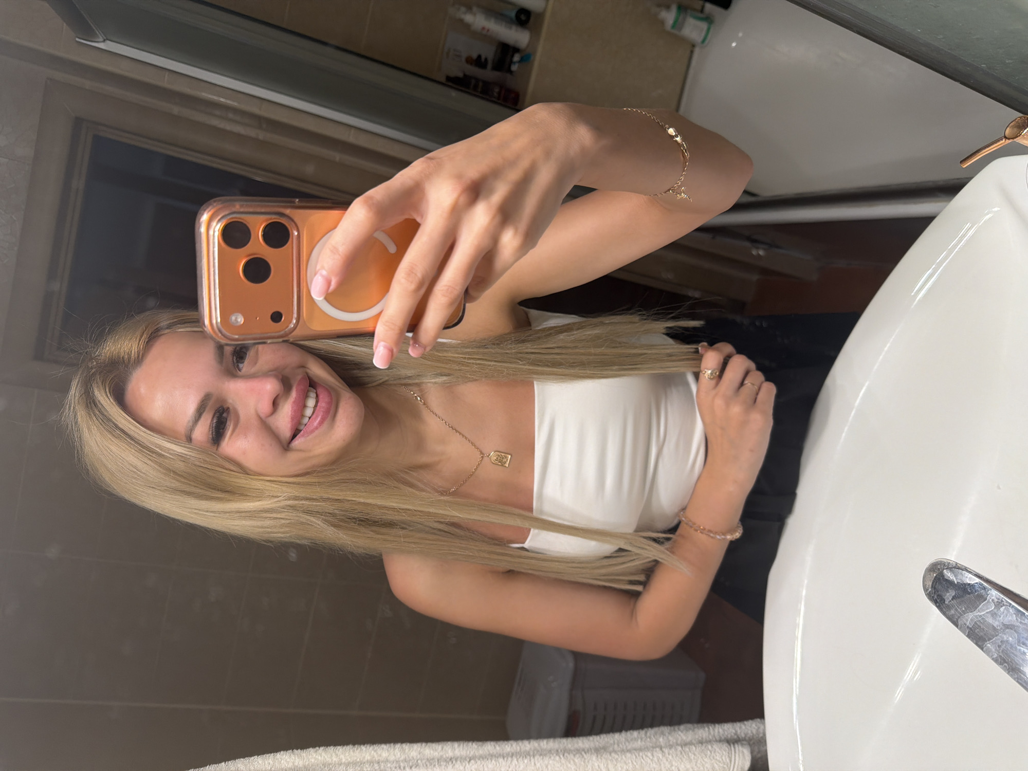 A young Caucasian woman with long blonde hair, wearing a white tank top and black pants, takes a mirror selfie in a bathroom. She is smiling, holding an orange smartphone in her right hand. She is wearing a gold necklace with a pendant and a bracelet on her left wrist. The bathroom appears to be in an apartment in Kyiv, Ukraine, with tiled walls and a white sink in the foreground.