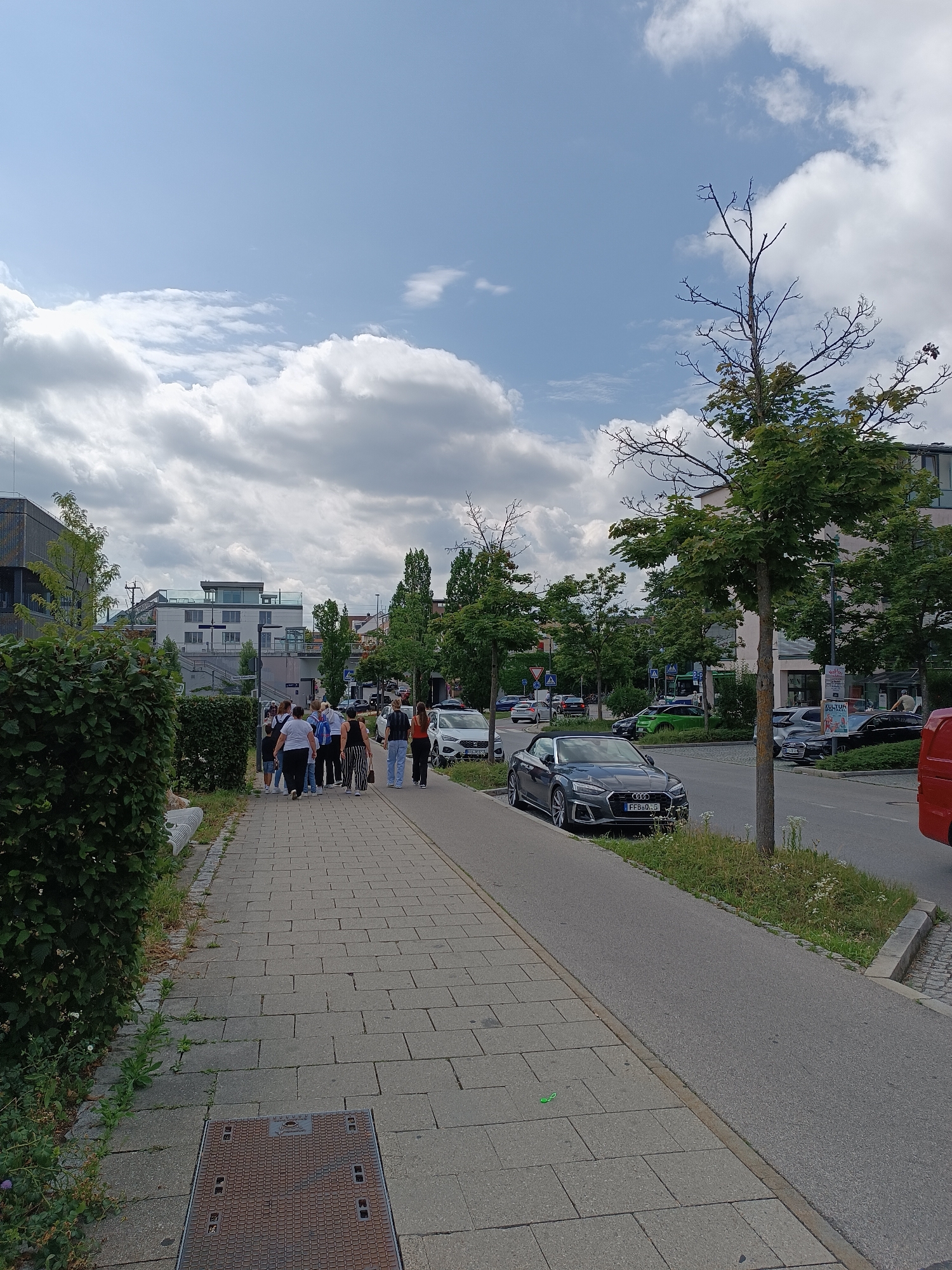 A group of approximately 10 people are walking on a paved pedestrian path next to a street in Germering, Germany. The path is bordered by a thick green hedge on the left and a road with parked cars and traffic on the right. Several cars are visible on the street, including a dark gray convertible, a white SUV, and a red van. Buildings and trees line the opposite side of the street. The sky is partly cloudy with a bright blue sky visible in some areas. The overall scene depicts a casual street view with pedestrians and vehicles under a daytime sky.