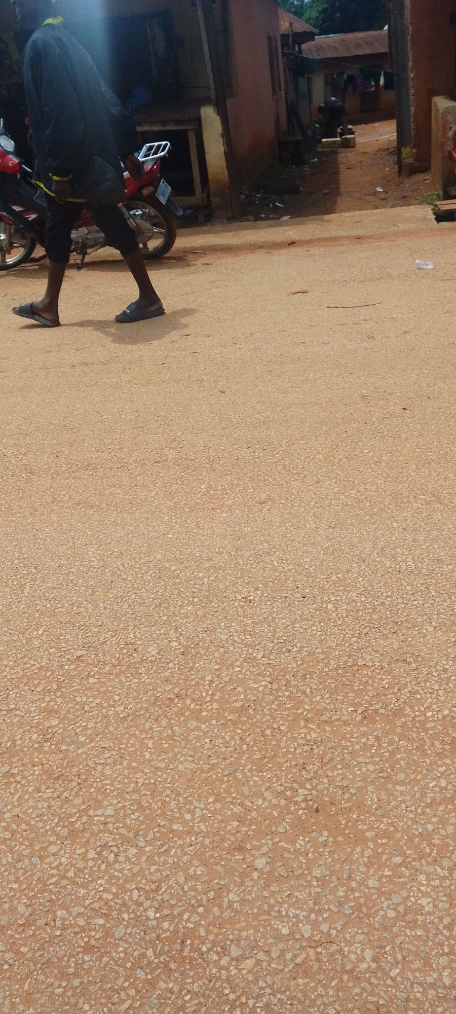 A person is walking on a dirt road in Baissa, Nigeria. The person is wearing a dark long-sleeved shirt, dark pants, and blue sandals. They are walking from left to right in the frame. A red motorcycle is parked to the left of the person. In the background, there are buildings and vegetation. The road is made of reddish-brown dirt and gravel. There are some small pieces of litter on the road. The lighting suggests it is daytime.