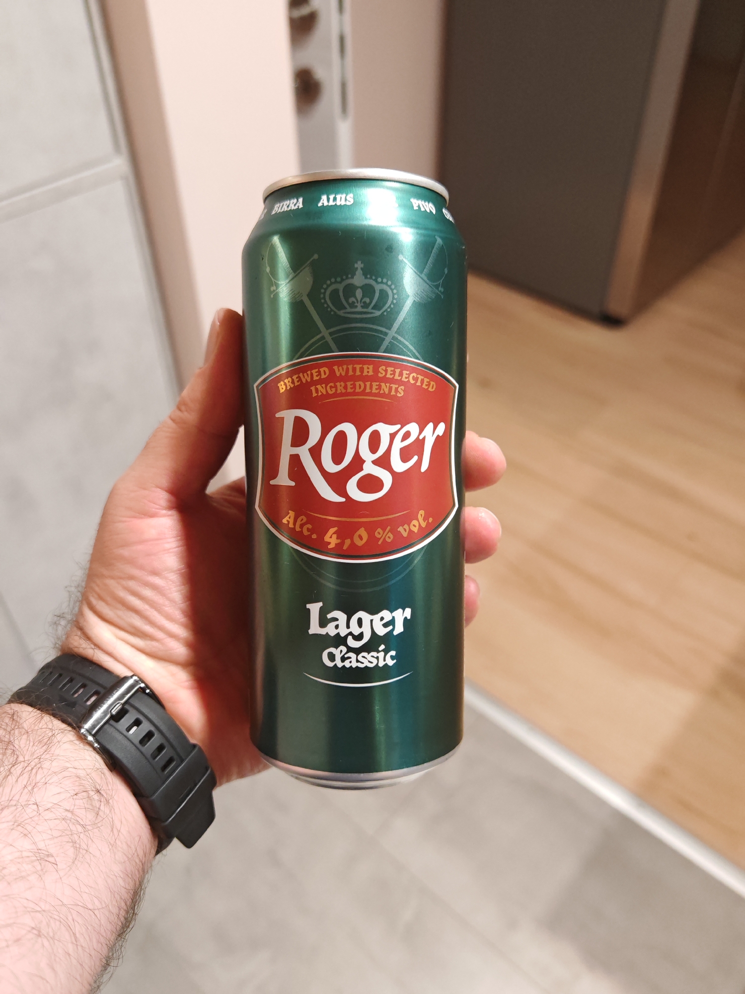 A hand, wearing a black watch, holds a green aluminum can of "Roger" lager beer. The can features a red label with the brand name "Roger" in large white lettering, surrounded by crossed swords and a crown. The text "BREWED WITH SELECTED INGREDIENTS" and "Alc. 4,0% vol." is also visible. The background appears to be an indoor setting with wooden flooring. The specific location in the city of Plovdiv, Bulgaria, is not discernible from the image.