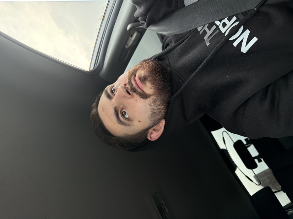 A male individual is depicted from the chest up, seated in what appears to be the driver's seat of a vehicle. He has short dark hair, a dark beard, and a mustache. His brown eyes are directed upwards and to the viewer's right. He wears a black hooded sweatshirt featuring the white "THE NORTH FACE" logo on the chest. A black seatbelt is positioned diagonally across his left shoulder and chest. The vehicle's interior includes a dark gray headliner and a partially open sunroof on the right side of the frame, revealing an overcast sky. A rearview mirror is partially visible in the lower left background. The specific location within the city of Zlatina, Bulgaria, cannot be determined from the visual evidence.