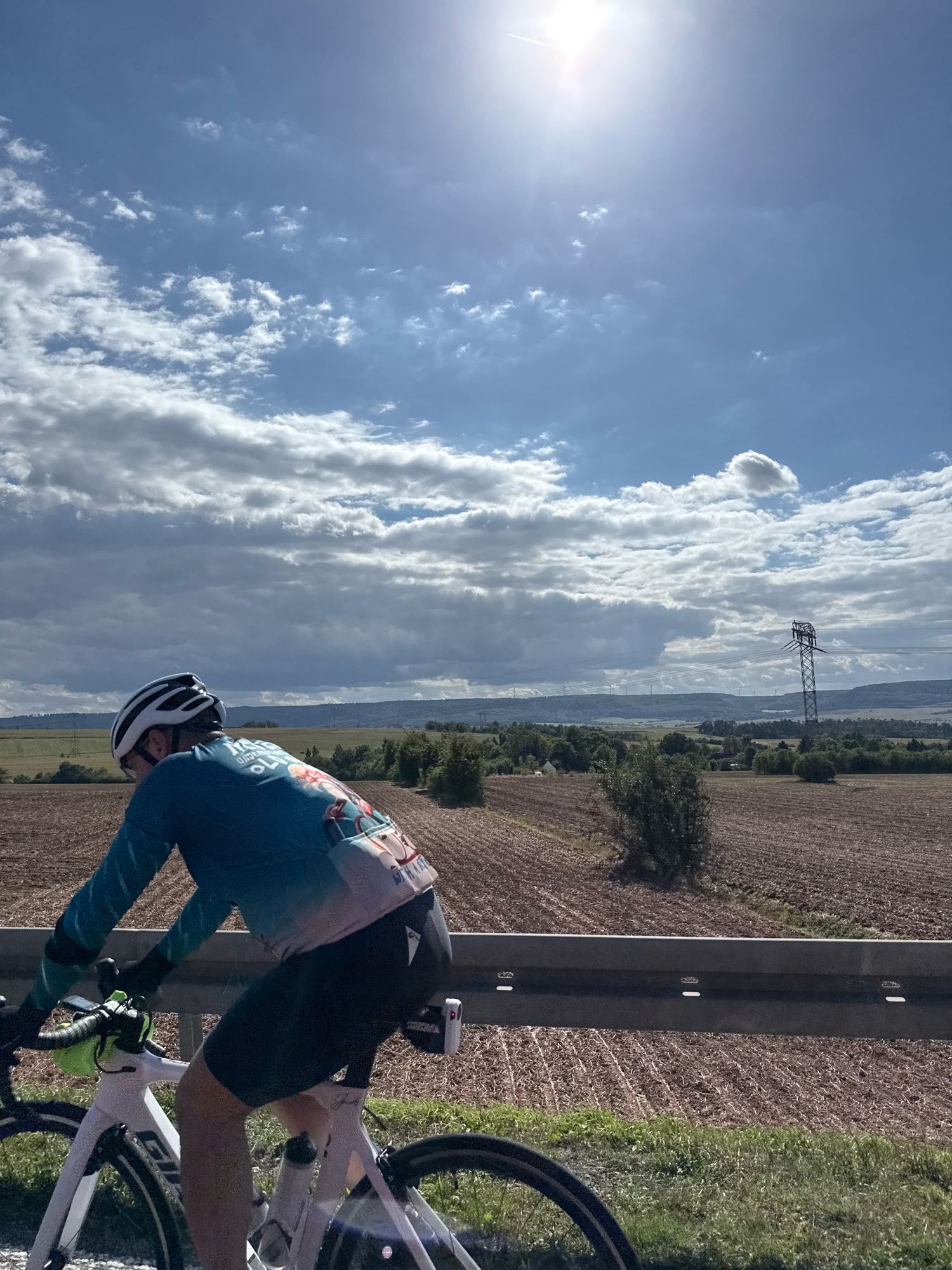 A person wearing a blue cycling jersey with a graphic design and a white helmet is riding a white bicycle on a road next to a guardrail. The bicycle has a green water bottle holder and a black saddlebag with a small white device attached. The background features a vast, open landscape with cultivated fields, trees, rolling hills, and a large power line tower. The sky is blue with scattered white clouds and a bright sun. The location is specified as Kleinfurra, Germany.