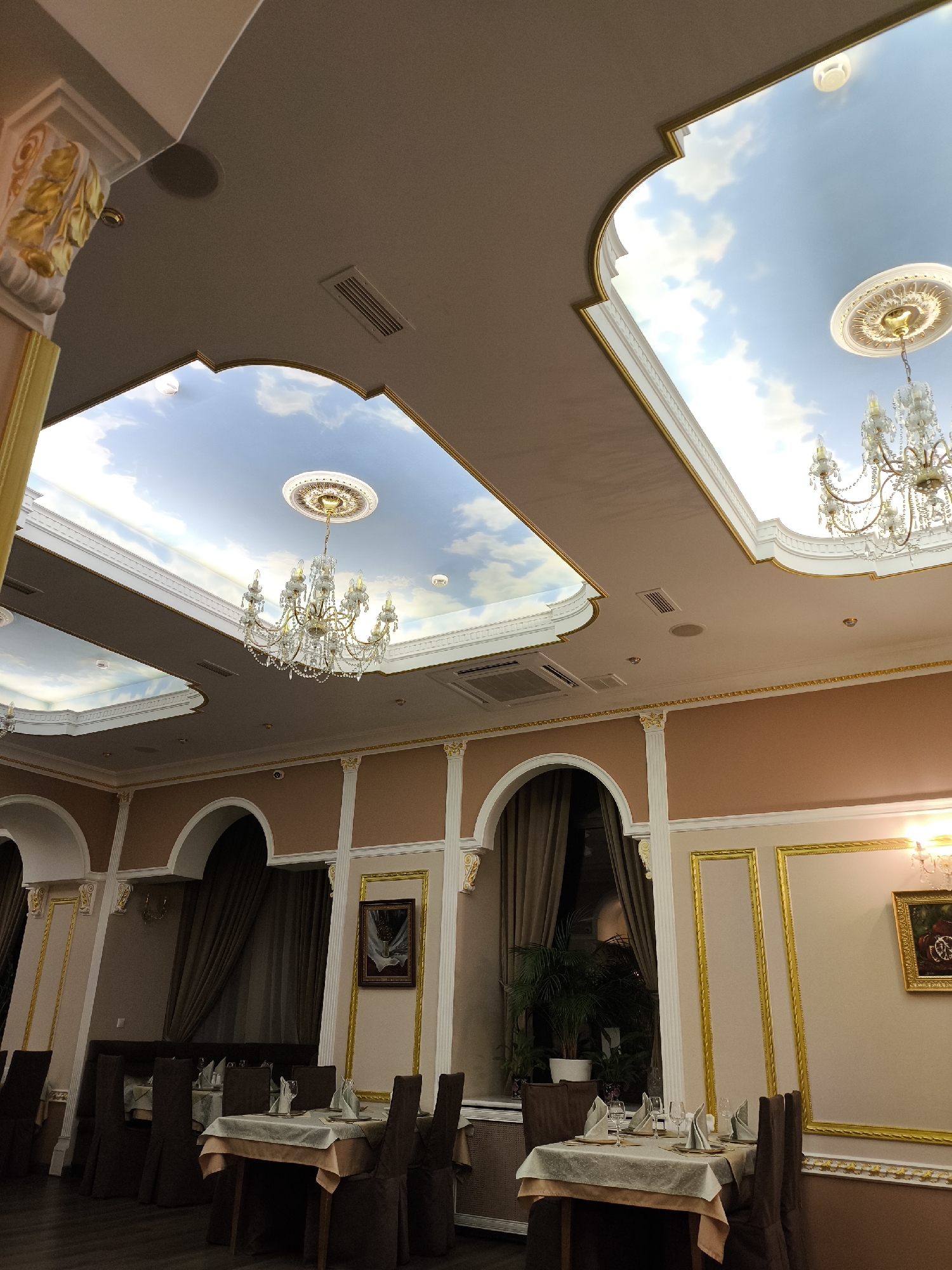 The image depicts the interior of a restaurant, likely located in Moscow, Russia, judging by the architectural style. The space features ornate decor, with a high ceiling decorated with trompe-l'oeil cloud paintings and elaborate chandeliers. The walls are adorned with gold-framed panels and artwork. Tables are set with tablecloths, glassware, and napkins, suggesting a formal dining setting. Chairs are upholstered in a brown fabric. The lighting is warm, creating an elegant atmosphere. No people are visible in the scene.