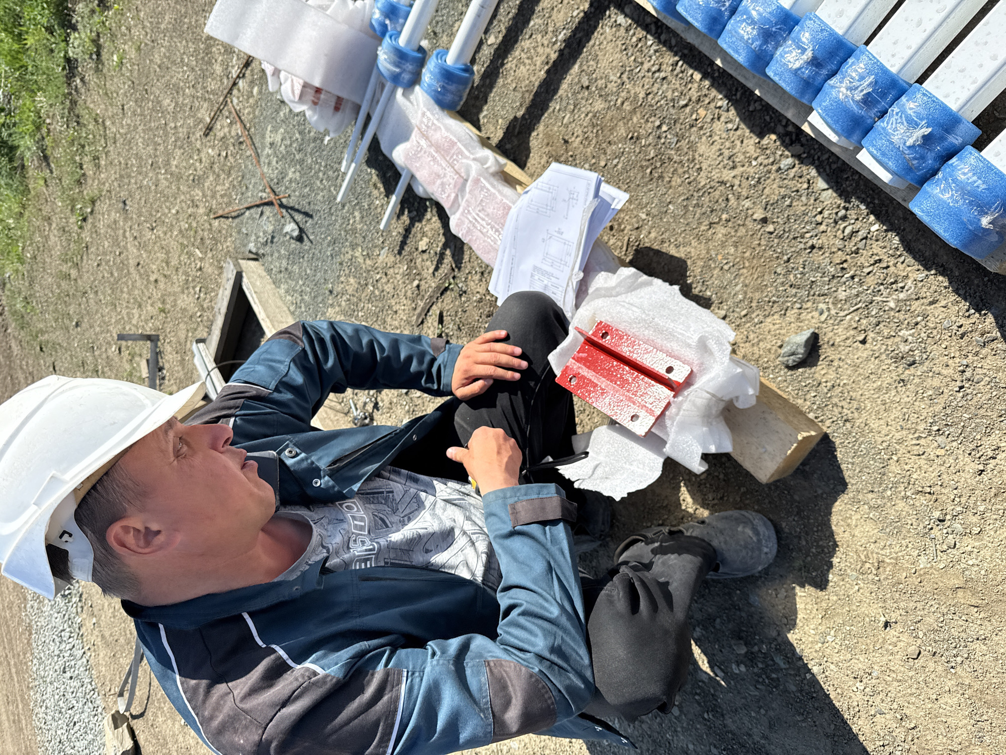 A male construction worker in Leninsk, Russia, wearing a white hard hat and blue work uniform, sits on a gravel surface. Next to him are construction materials, including red painted metal parts, white metal rods bundled with blue protective wraps, white paper, and a piece of wood. He appears to be inspecting or preparing for a construction task.
