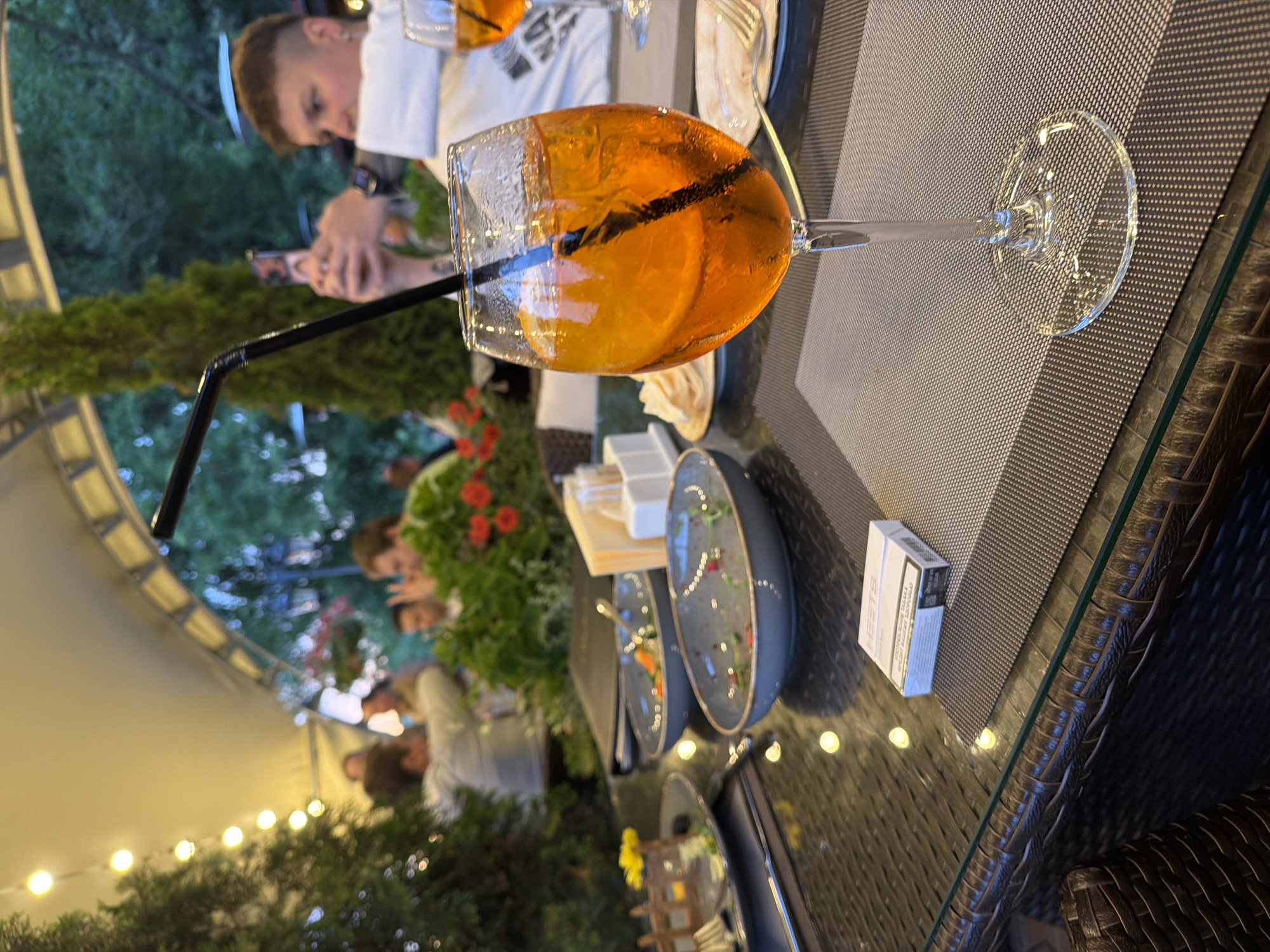 The image depicts an outdoor seating area of a restaurant or cafe, likely in Izmailovo, Russia, as indicated by the specific context of the prompt. The scene is set at a table with a glass-top surface, adorned with woven placemats. In the foreground, a tall stemmed glass filled with an orange-colored beverage, garnished with a slice of orange and a black straw, is prominently featured. Beside it, a pack of HEETS cigarettes rests on a placemat.
The table is also set with several used dishes, suggesting a meal has been or is being consumed. One plate contains remnants of food and scattered greenery. Another deeper bowl holds a small amount of salad or vegetables. A cutlery set and a small plate with what appears to be bread or crackers are also visible.
In the background, blurred figures of people are seen seated at tables under a white tent-like canopy, illuminated by a string of lights. Trees and foliage frame the outdoor setting. One person in the mid-ground is holding up a mobile phone, possibly taking a photograph. The overall atmosphere suggests a relaxed, social gathering during daylight hours, possibly late afternoon or early evening, given the lighting.