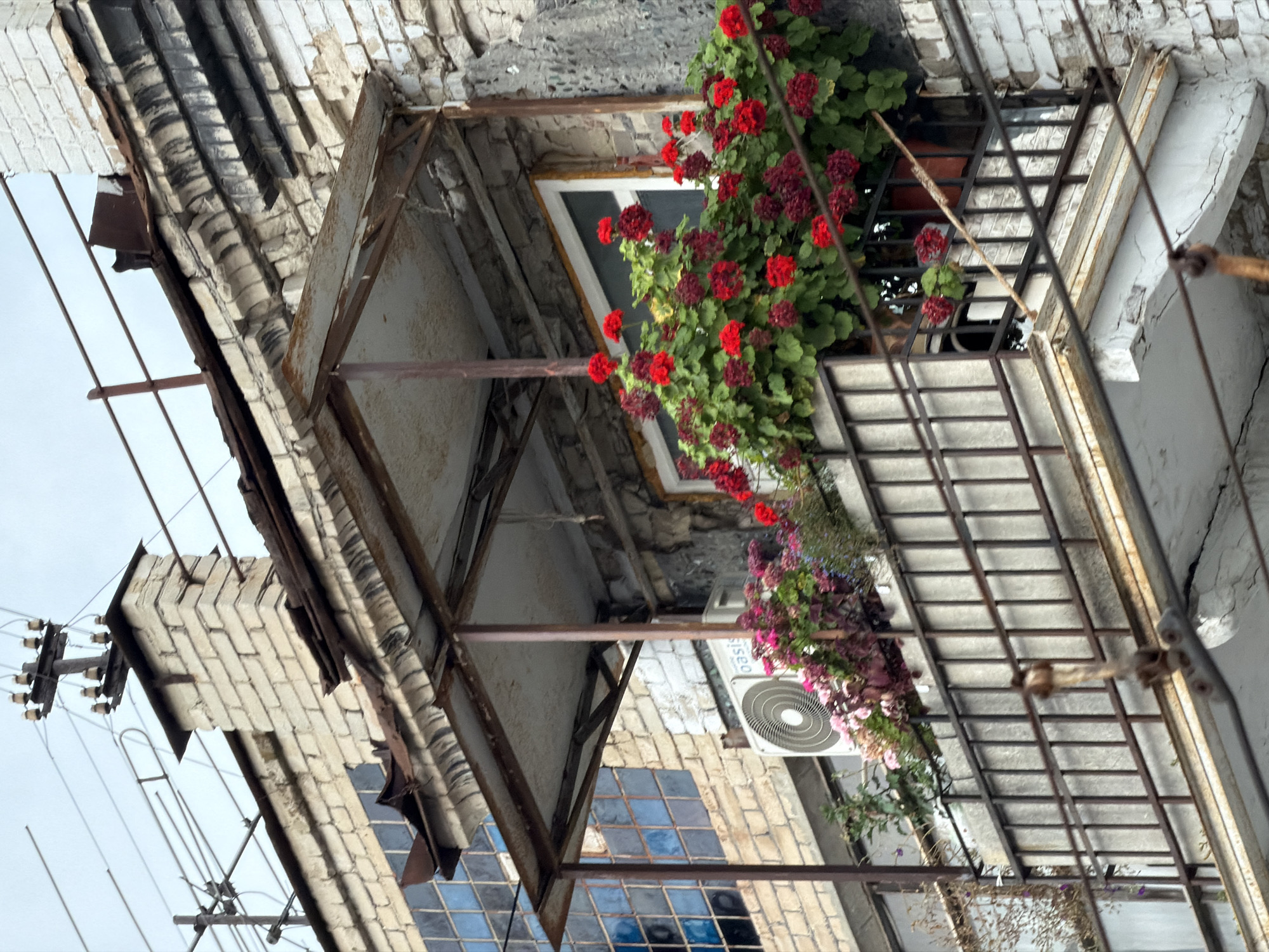 The image shows a detail of a dilapidated building's facade, likely an apartment block, in Saratov, Russia. The foreground features a rusty metal balcony railing with a concrete floor. Above the balcony, a window is framed by lush red and dark pink flowers in planter boxes. An air conditioning unit is visible to the left of the window. The building's exterior is constructed of weathered white bricks, with visible signs of age and neglect, including peeling paint and staining. A rudimentary metal awning juts out above the balcony. Further up, a brick chimney is visible against a cloudy sky, with electrical wires and poles attached to the rooftop. The overall impression is one of urban decay with touches of natural beauty from the flowers.
