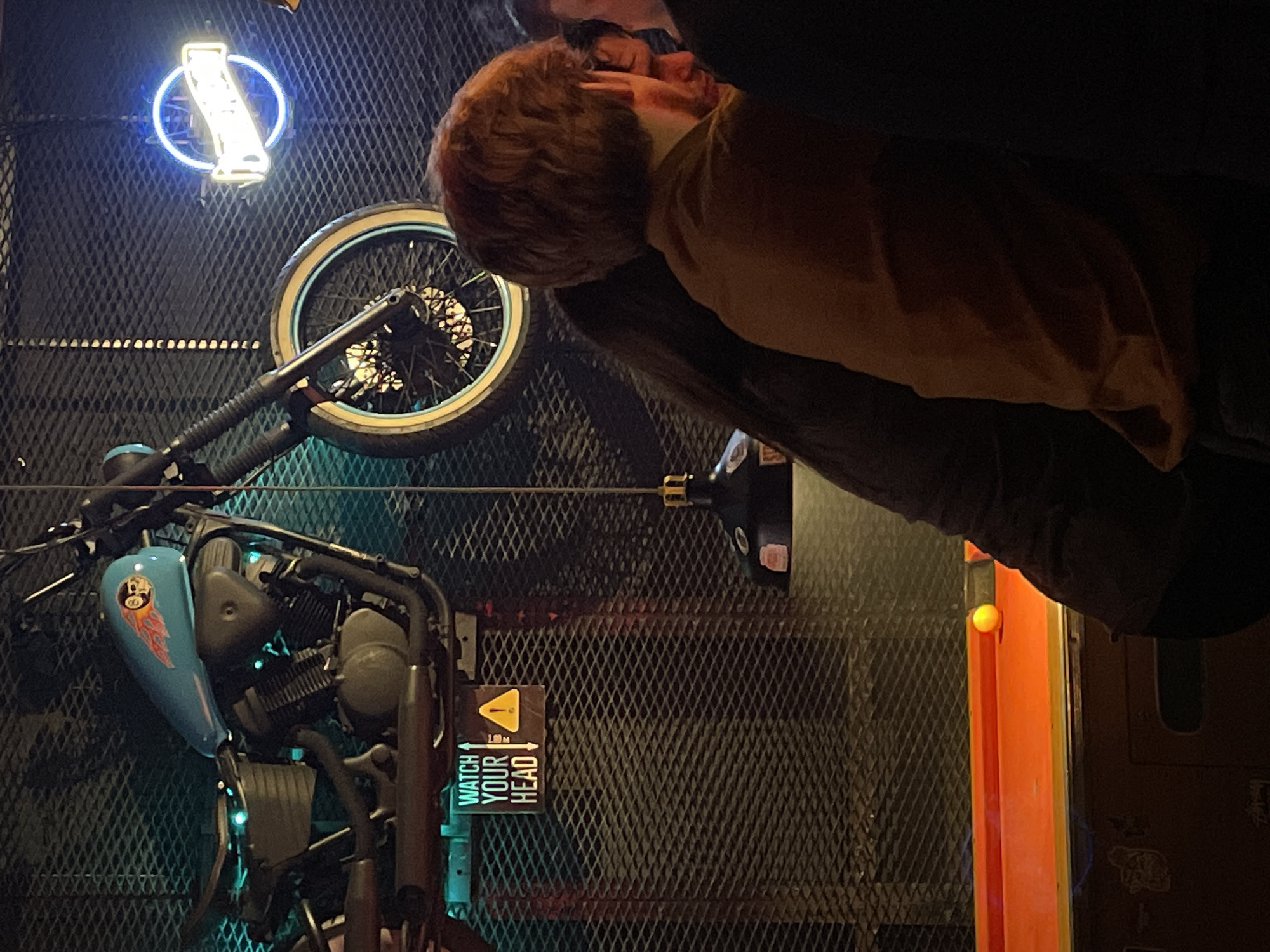 A motorcycle is mounted on a black mesh wall. Above the motorcycle is a wheel and a neon sign. A person with red hair is near the wall. There is a "watch your head" sign. An orange beam is near the mesh. The image was taken in Belgrade, Serbia.