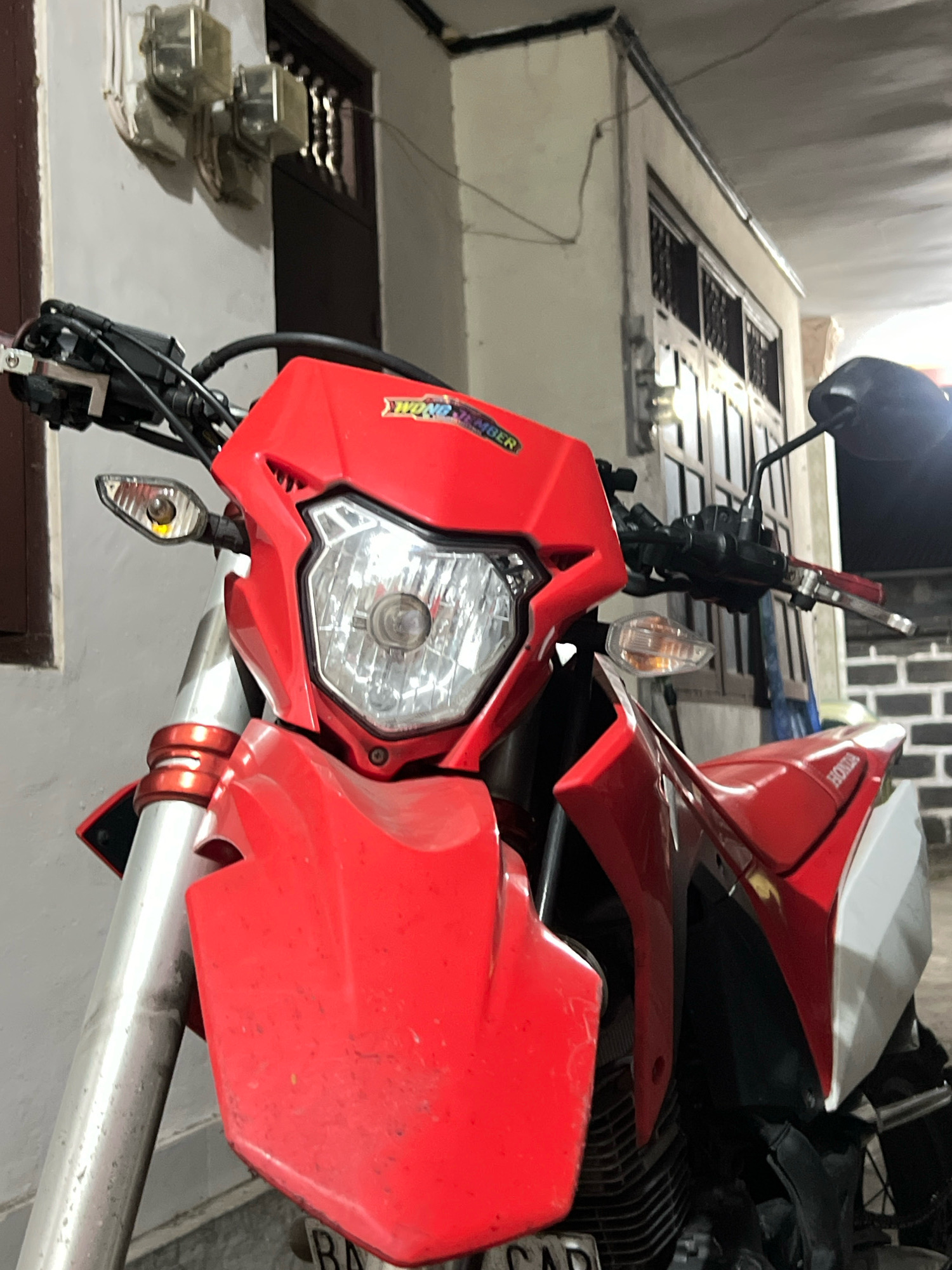 A red and white Honda dirt bike is parked in front of a building. The bike has a prominent red front fender and headlight assembly. The handlebar area, mirrors, and part of the seat are also visible. The bike is positioned close to a wall on the left side of the frame, which has some electrical meters attached. The background shows a building entrance with a dark-colored door and barred windows. The scene appears to be an outdoor urban setting, likely a residential or commercial area in Denpasar, Indonesia, given the architectural style and the presence of electrical meters commonly found in the region. There are no people visible in the image.