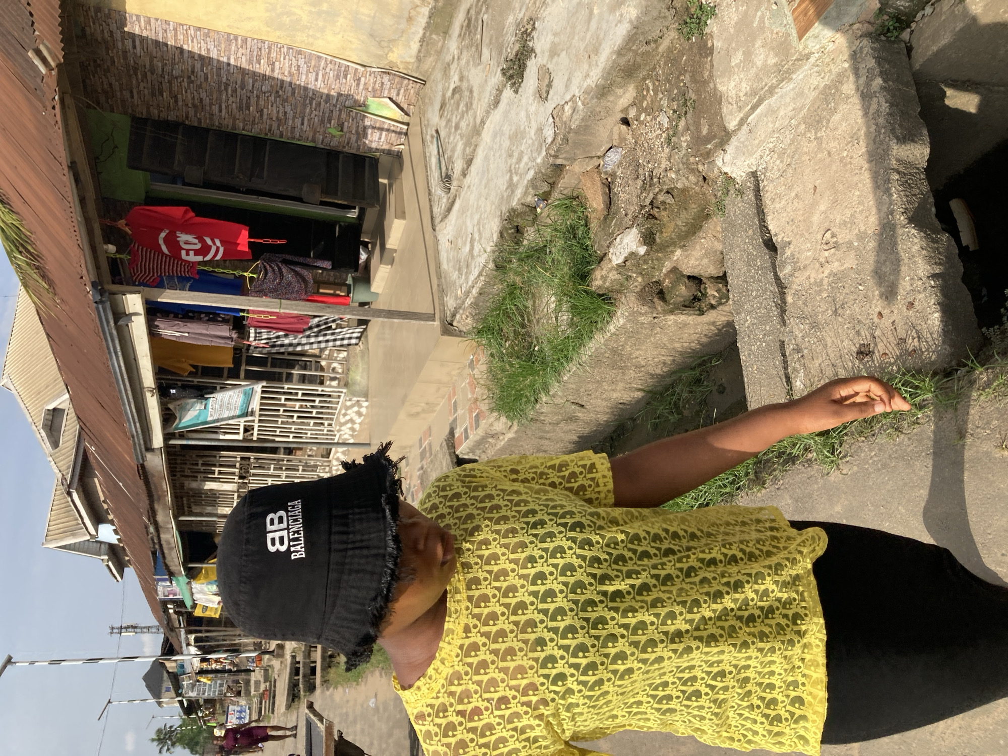 A person wearing a black Balenciaga bucket hat and a yellow Dior print top is standing outside. The location is likely Aba, Nigeria, based on the architectural style of the buildings in the background. The buildings have corrugated metal roofs, and the area appears to be a commercial district with shops. Clothing is displayed for sale outside one of the shops. Concrete surfaces are visible near the person, with some grass and weeds growing along the edges.