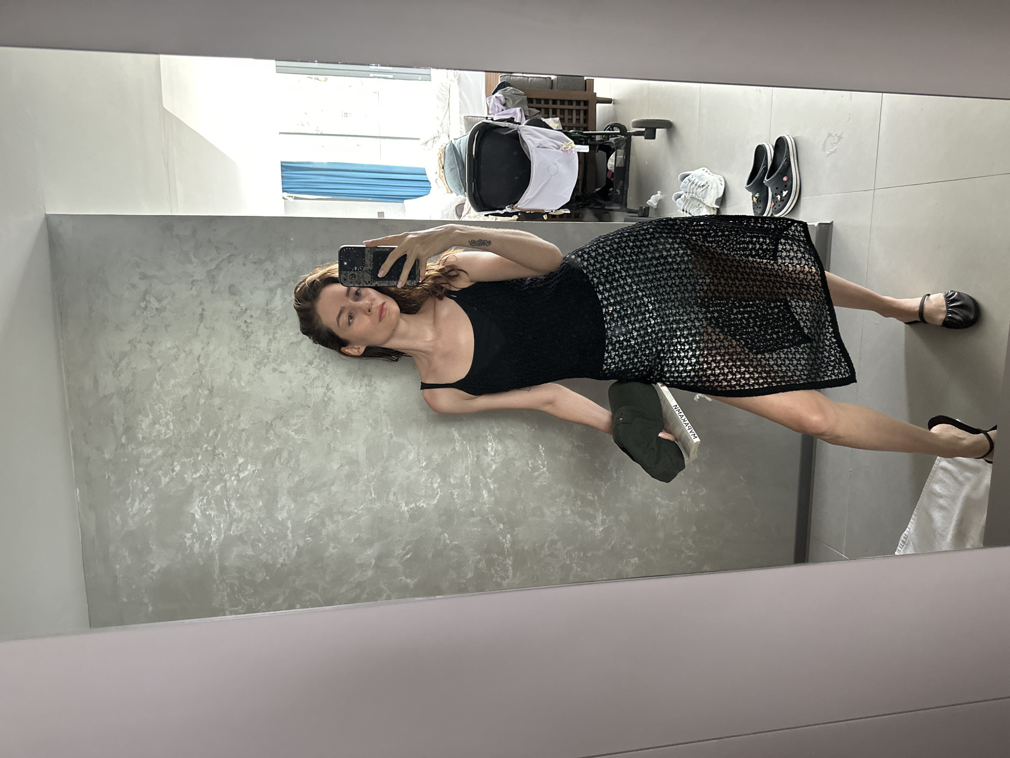 A woman is taking a selfie in a mirror. She is wearing a black tank top and a black mesh skirt. She is holding a phone in her left hand. Her right hand is holding a green shoe. There is a stroller in the background. The scene is located in Choeng Thale, Thailand.