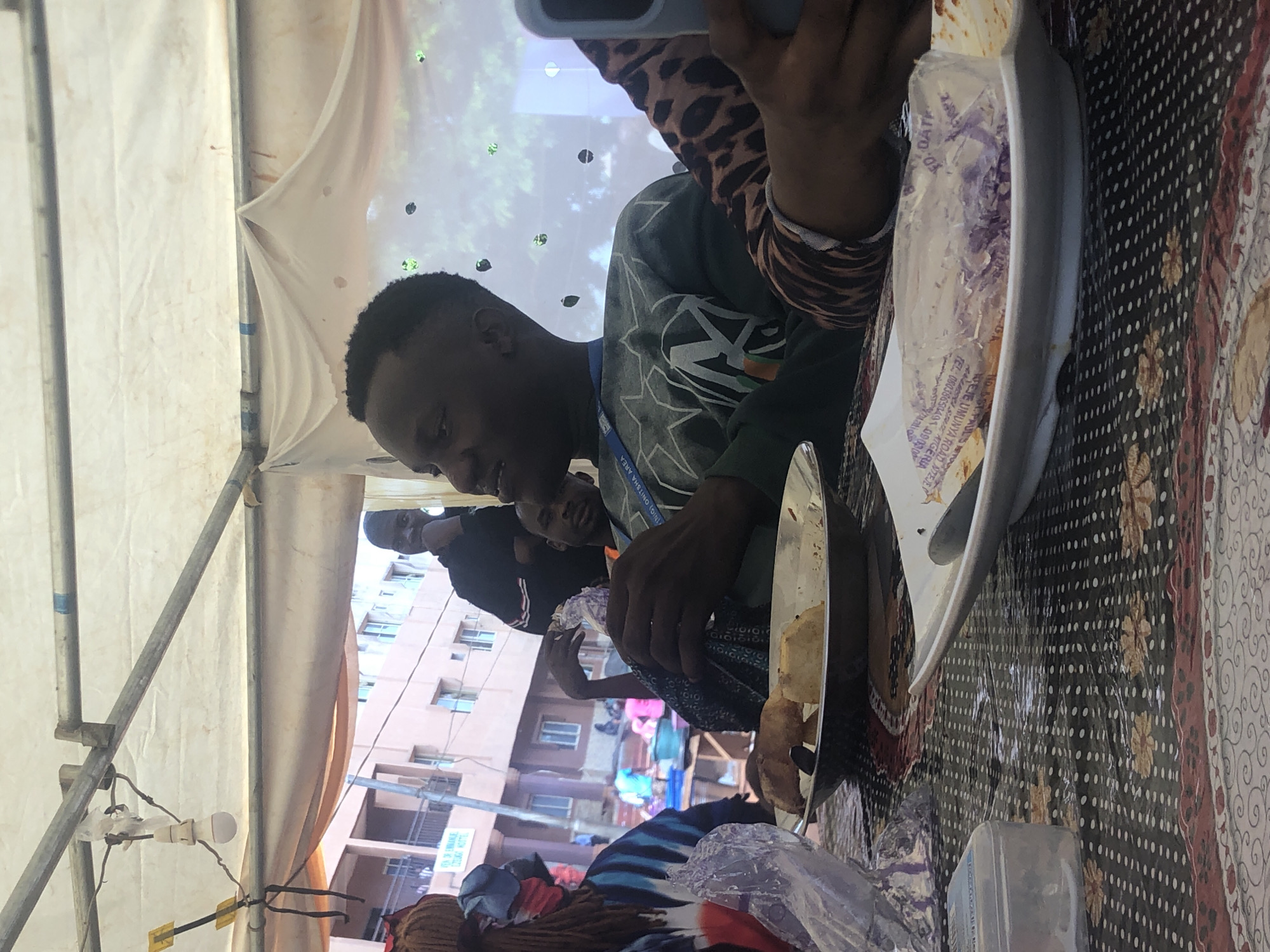 The image shows a group of people eating a meal under a tent-like structure. A man in a green sweatshirt with an orange lanyard around his neck is the main focus, seemingly engaged in eating. He is seated at a table covered with a patterned tablecloth. Another person with a leopard print sleeve is partly visible next to him, holding what appears to be a phone or tablet. In the background, other individuals are visible, along with buildings, hinting at an urban environment. A building with a sign is visible with words: ST PAULS TO, WEJE UMUNYA ROAD NIEJE, ANAMERA SATGERIA. From the text, it is determined the location is near Weje Umunya Road in Nteje, Nigeria.