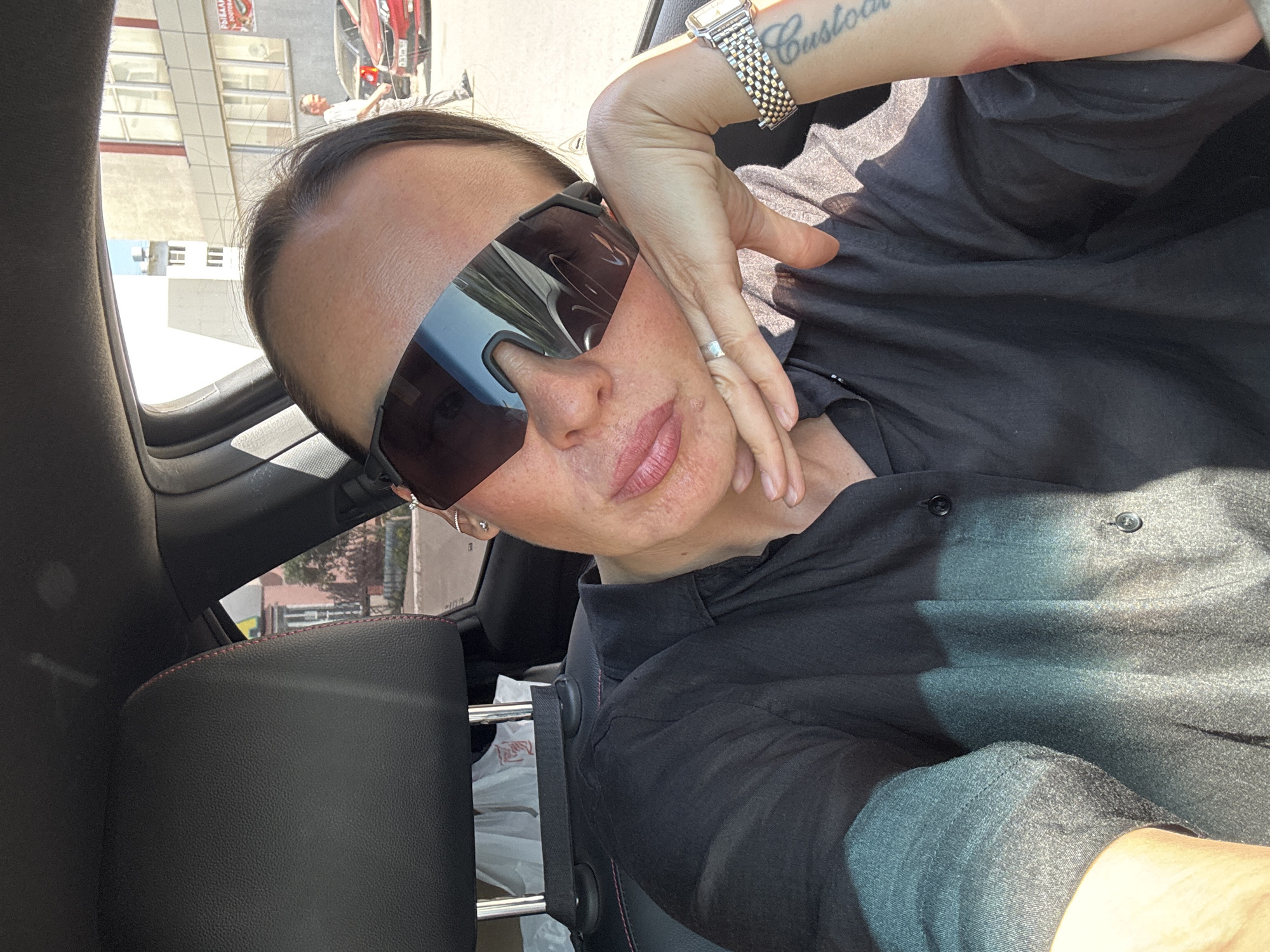 A person with sunglasses sits in a car, wearing a dark shirt and a wristwatch. The person's left hand is near their face. Reflected in the sunglasses are buildings and possibly a red car. Outside the car window are city buildings. The location is Chaikovsky, Russia. The car appears to be parked.