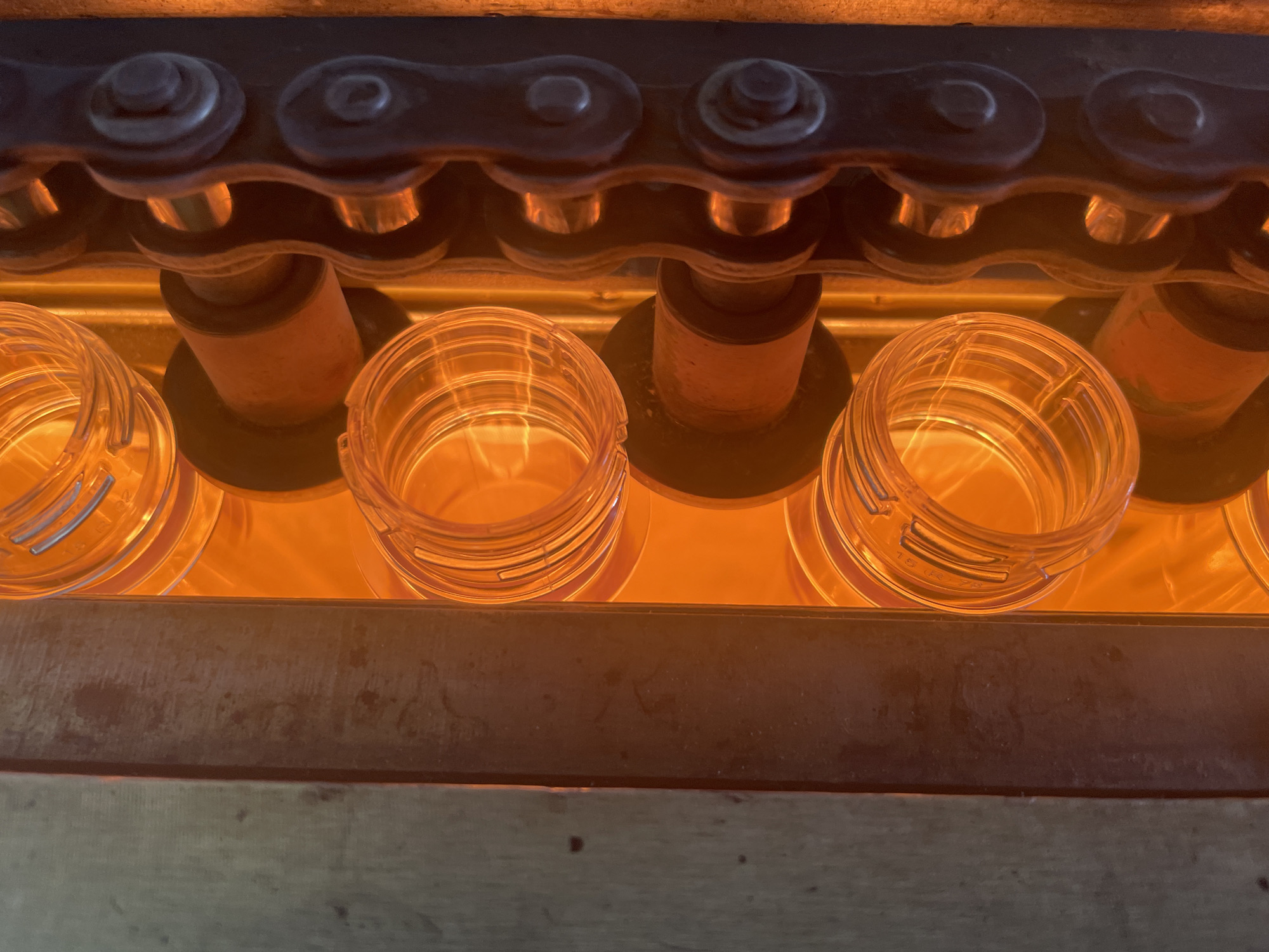 A close-up, vertical shot captures clear plastic bottle preforms being heated and transported on a conveyor chain. The preforms are cylindrical with visible ridges and are glowing orange from intense heat. They are positioned vertically and appear to be moving from left to right. A dark metal chain with circular links is visible on the right side of the frame, guiding the preforms. The background consists of a blurry orange glow, indicating a heat source or an oven. To the left, a textured, dark grey surface, possibly metal or wood, forms the edge of the machinery. The scene appears to be an industrial setting, likely a manufacturing plant for plastic bottles. The location is identified as the city of Tervolovo, Russia. There are no people visible in the image.