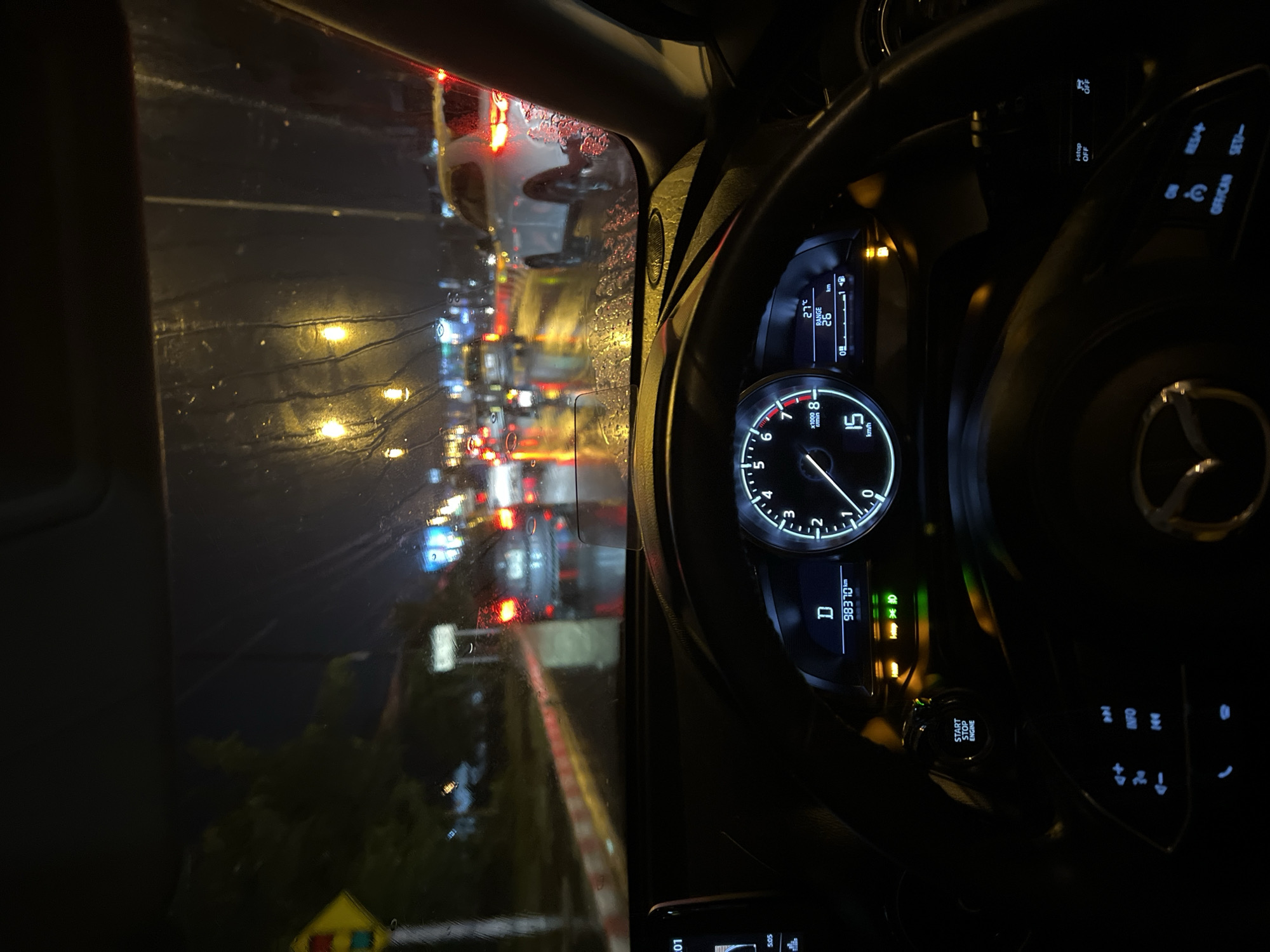 Here is a description of the image:
The image shows the interior of a car, specifically the dashboard and steering wheel, at night. The car is in traffic, as seen through the rain-streaked windshield, with numerous other vehicles visible ahead, their lights blurred by the wet glass.
The car's dashboard is illuminated, displaying the speedometer indicating a speed of 15 km/h. The tachometer reads just above 0. The odometer displays "98370km". The dashboard also shows an ambient temperature of 27 degrees Celsius and a range of 66 km.
The steering wheel features various control buttons and the car's logo, which is the Mazda logo. Outside, through the windshield, the scene is dark with lights from other vehicles and streetlights reflecting on the wet road and the rain-covered windshield. The traffic signal is visible.