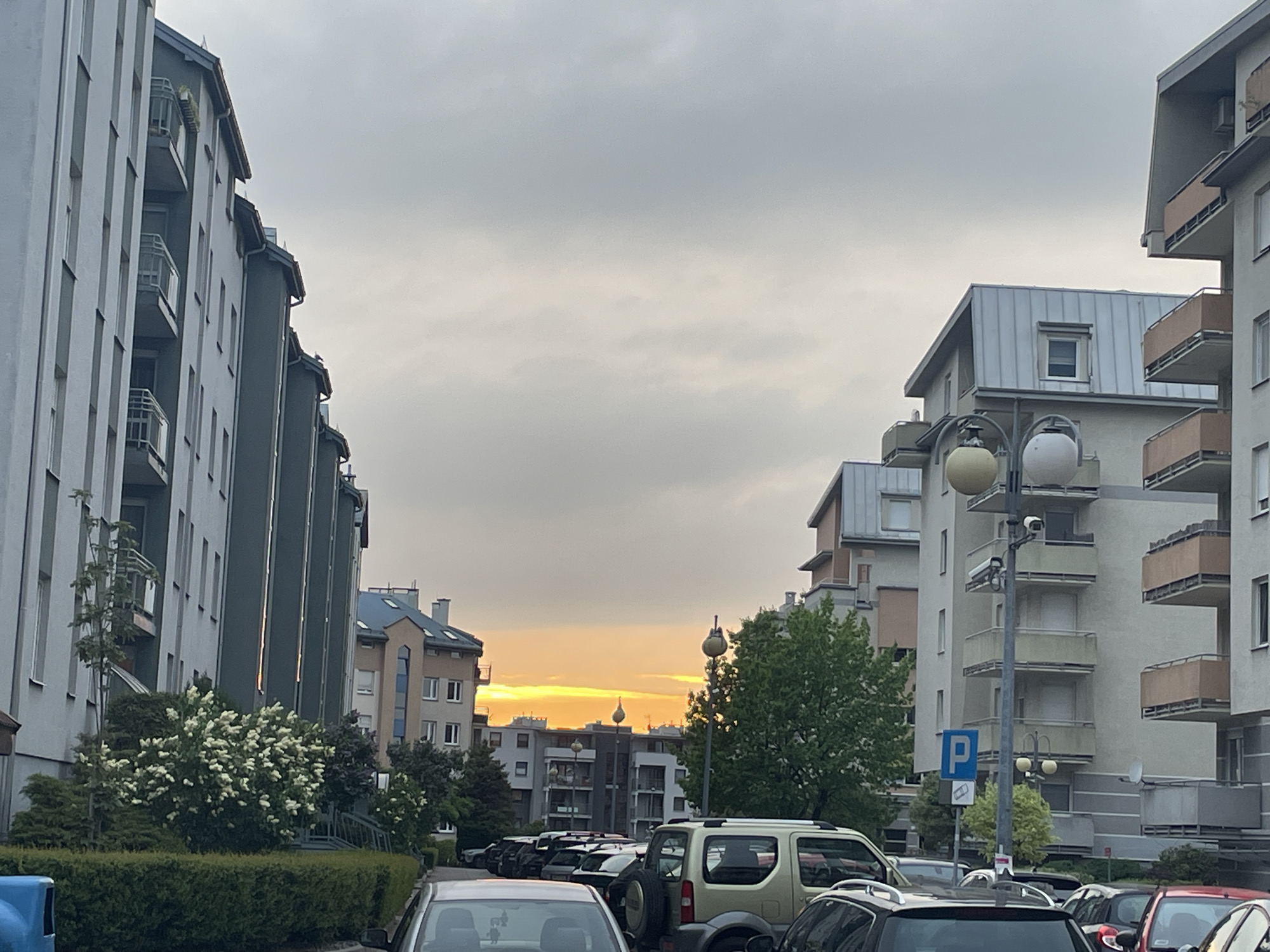 The image depicts a street scene in an urban environment, possibly a residential area. Rows of parked cars fill the foreground, partially obscuring the lower portions of buildings in the background. The buildings on either side of the street are multi-story apartment complexes or residential buildings, with varying architectural styles and balconies. The sky is overcast, with a hint of orange or yellow visible on the horizon, suggesting a sunrise or sunset. Streetlights are visible along the street, and some greenery, including trees and bushes, is present. A "P" sign indicates a parking area. The location is likely in Unknown, Unknown.
