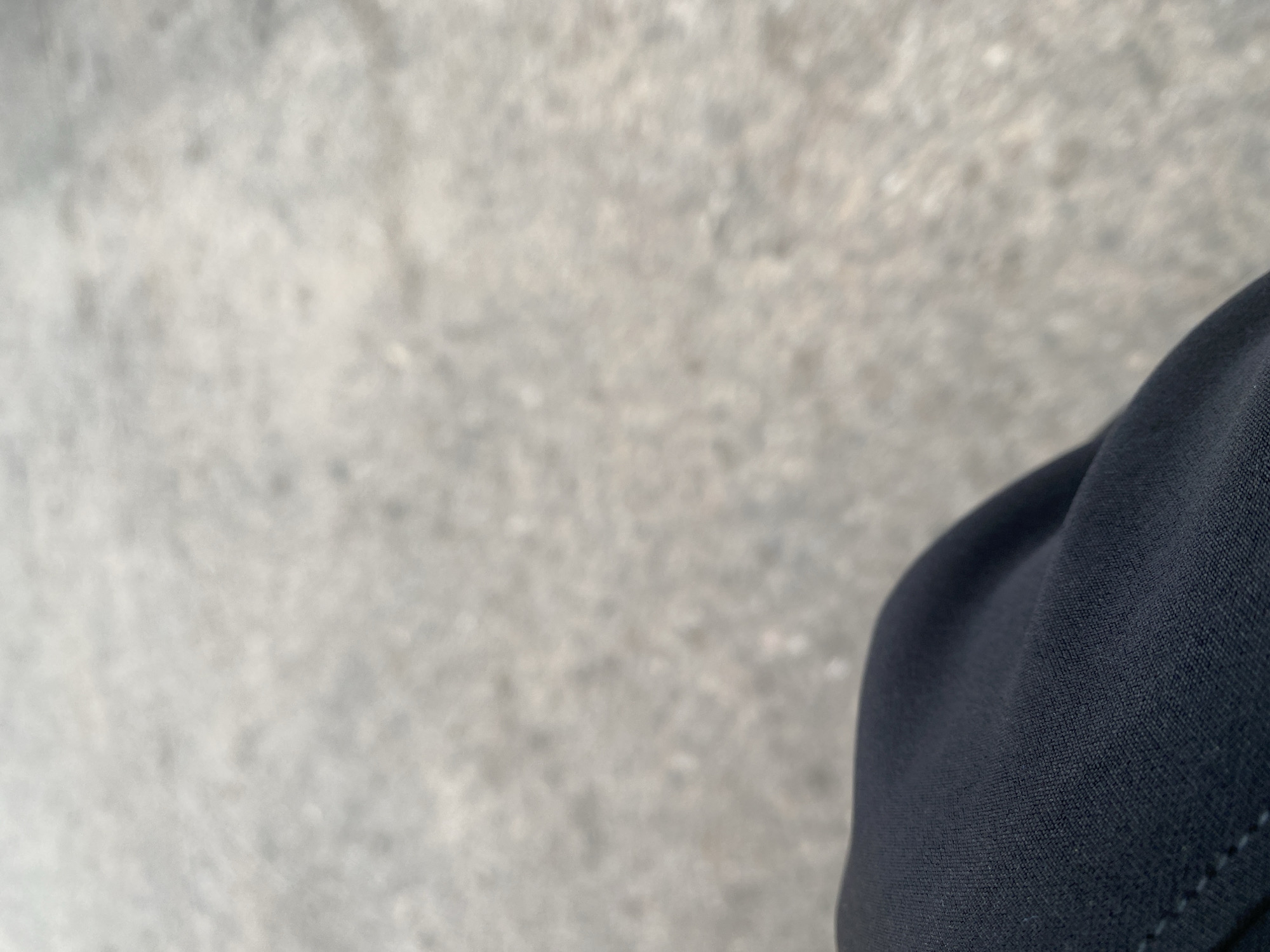 The image shows a close-up view of dark fabric, likely clothing, in the foreground. The fabric is textured and appears to be a deep navy blue or black. The background is out of focus and shows a mottled, greyish-brown surface that resembles pavement or a textured ground. There are no discernible people, objects, actions, or interactions visible in the scene. The image does not provide any visual information that would allow for the determination of its location within Jalingo, Nigeria.