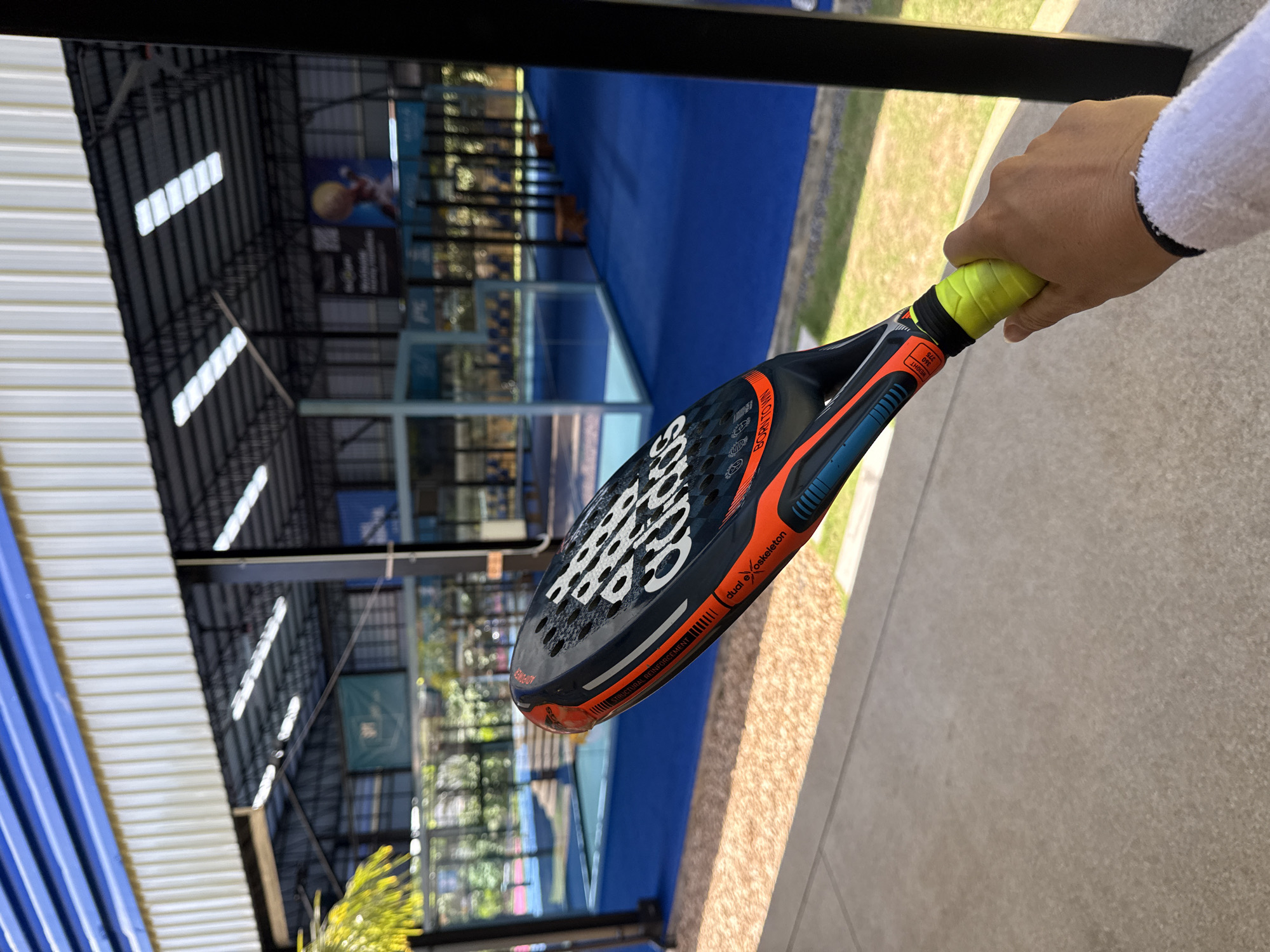 A hand grips a yellow tennis racket handle wrapped in a white towel sleeve, with a paddle tennis racket held in front of it. The racket is black with orange and blue accents, and white "adidas" lettering. In the background, a bright blue paddle tennis court with glass walls is visible under a covered roof structure. The scene appears to be outdoors at a sports facility. There is no indication of the location being in Ban Rang Rak, Thailand.