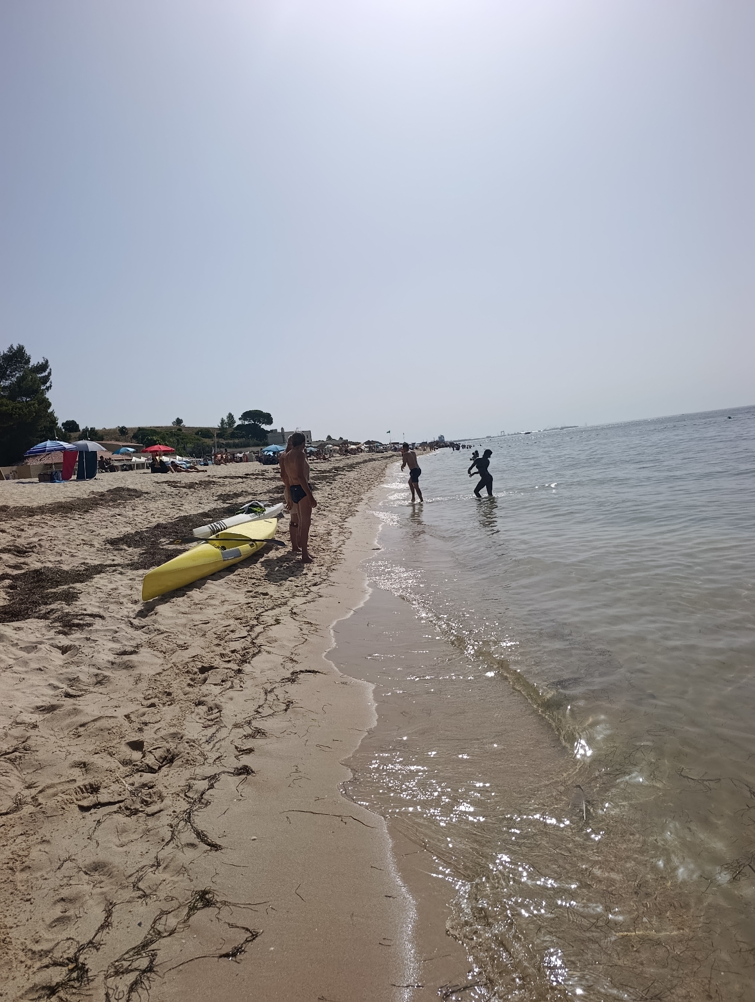 The image depicts a sunny day at a beach with a calm sea. In the foreground, two kayaks are resting on the sand, a yellow one and a white one. Two people, a man and a woman, are standing near the kayaks, looking towards the sea. Further down the beach, several beach umbrellas are set up, providing shade for beachgoers. Some people are visible relaxing on the sand or walking along the shore. In the water, three people are wading or playing near the shoreline. The sea is clear with gentle waves lapping at the sand. The background shows a line of trees and some buildings, suggesting a developed coastal area. There is no specific indication that this scene is located in Setúbal, Portugal.