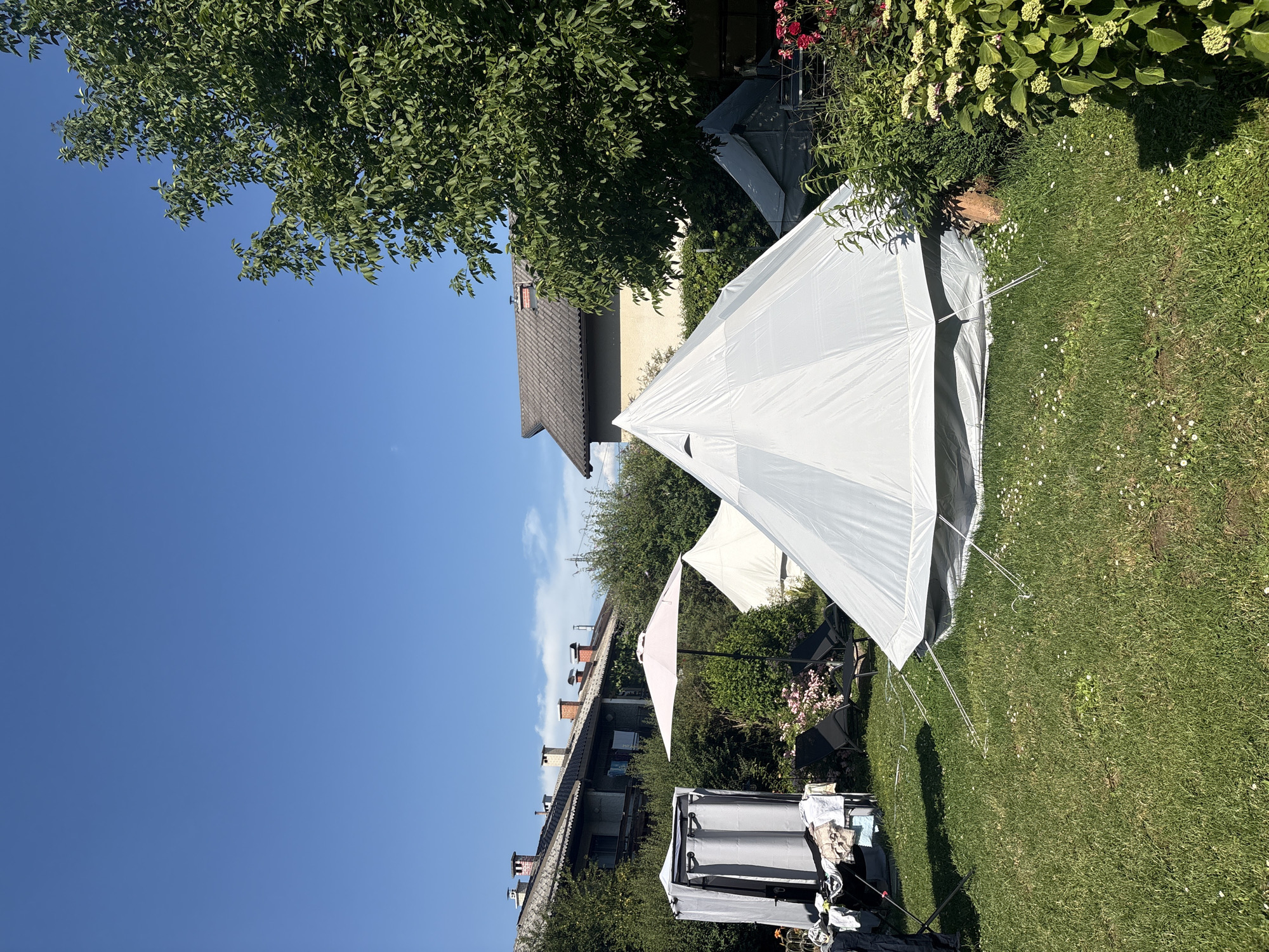 An outdoor shot shows a garden with several tents set up. The main tent is a large, light-colored bell tent in the foreground. To the left of the bell tent, there's a smaller white tent. Further back, another grey tent is visible. A pink umbrella stands near the smaller white tent, and two black lounge chairs are positioned on the grass. On the far left, a portable changing tent is set up, with clothes draped on a drying rack next to it. Behind the tents and garden, residential buildings with tiled roofs and multiple chimneys are visible. A large tree with green leaves occupies the right side of the frame, casting a shadow on the grass. The sky is clear and blue.