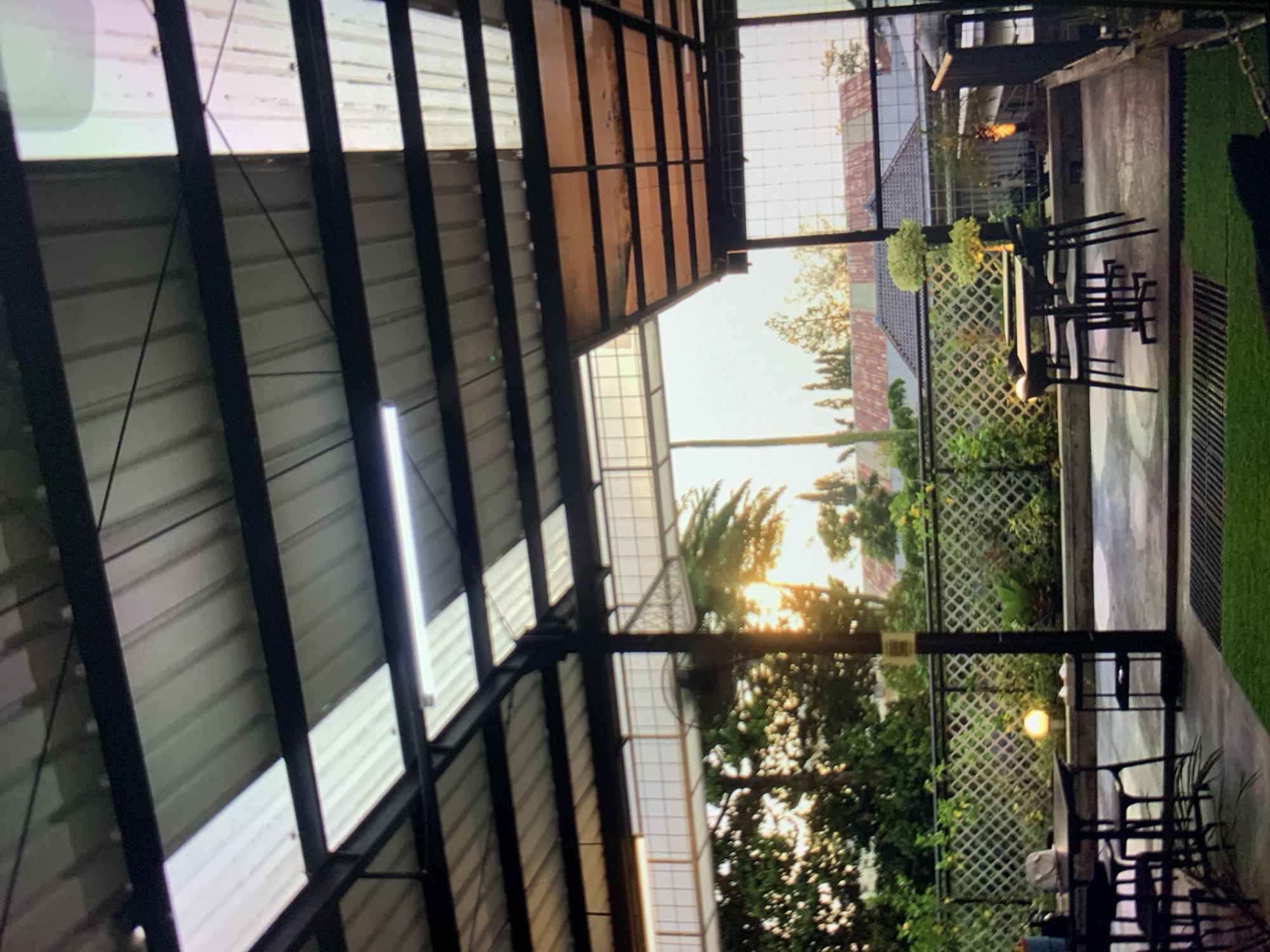 The image depicts an outdoor seating area of a cafe or restaurant, likely located in Ciamis, Indonesia, given the context. The scene is captured during sunset, with the warm glow of the sun filtering through the translucent roof panels and surrounding trees. In the foreground, several tables and chairs are arranged on a concrete patio, with a patch of artificial grass visible. A black metal fence separates the seating area from a garden with various plants. In the background, residential buildings with tiled roofs can be seen amidst lush greenery. A large fan is mounted on a pole, and a streetlamp casts a soft light on the garden. The overall ambiance suggests a peaceful evening setting for outdoor dining or relaxation.