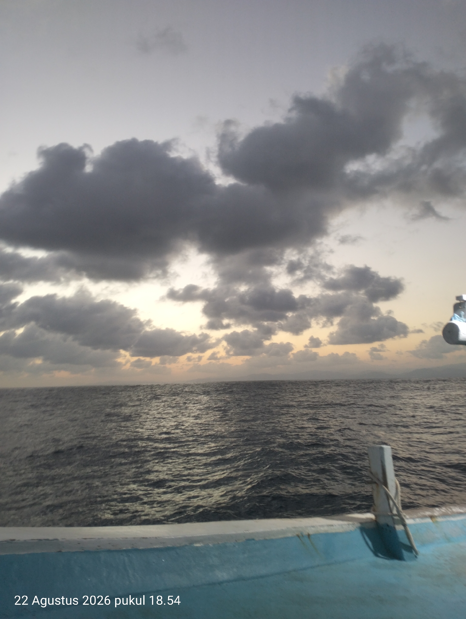 This is a wide shot of a body of water under a cloudy sky at dusk. The camera is at water level, looking out over the ocean. The water is dark and choppy. In the foreground, the edge of a light blue boat is visible, with a white cleat and rope. In the distance, a hazy landmass is visible on the horizon. The sky is filled with dark, dramatic clouds, with hints of orange and yellow light breaking through near the horizon. The timestamp at the bottom of the image reads "22 Agustus 2026 pukul 18.54". The location is not specified.
