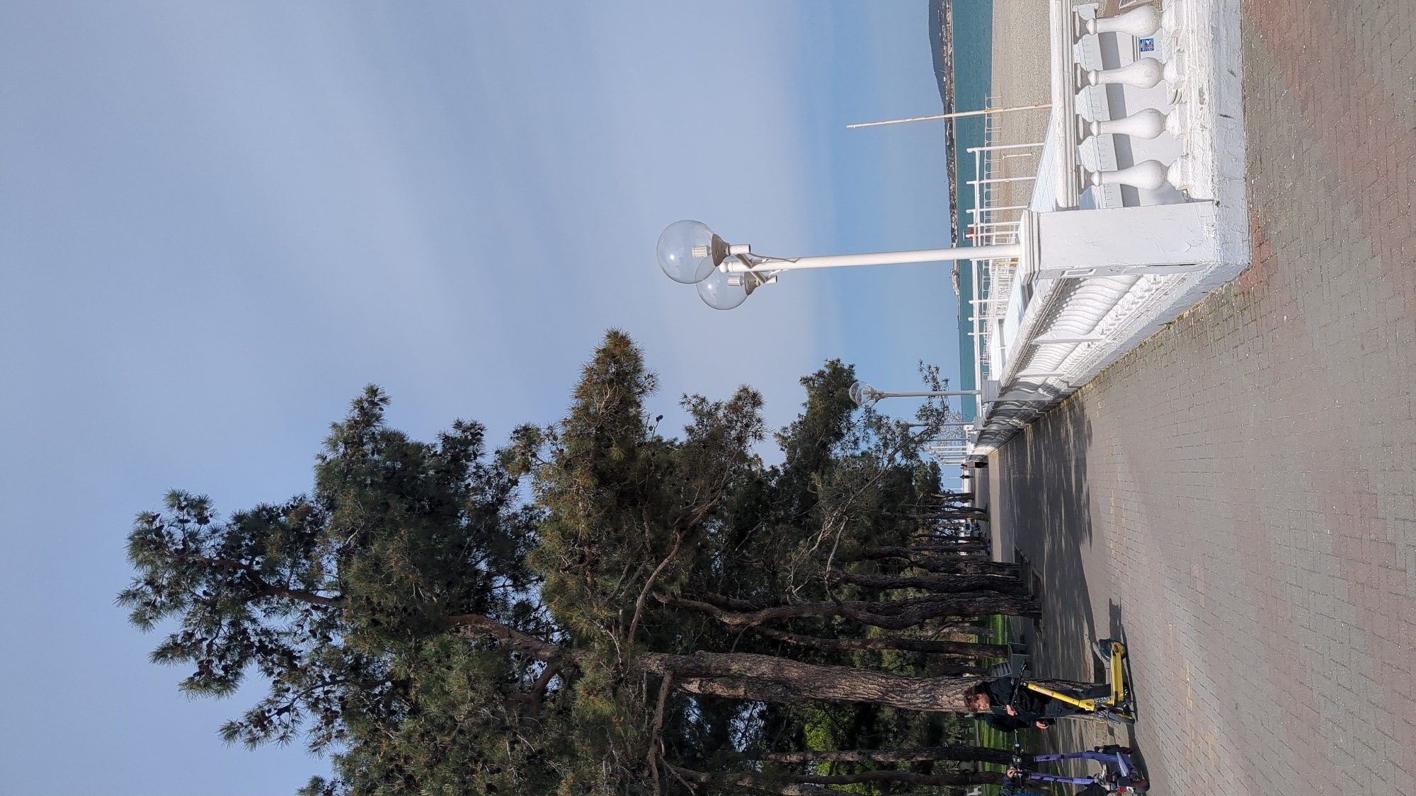 This image depicts a sunny day on a seaside promenade in Gelendzhik, Russia. A wide, paved walkway, composed of reddish-brown and gray pavers, stretches from the foreground into the distance, running parallel to the coastline.
On the left side of the promenade, a dense line of mature evergreen trees, likely pines, stands tall, their dark green foliage contrasting with the light blue sky. These trees cast dappled shadows across the pathway. On the right, a pristine white balustrade with decorative pillars acts as a barrier, overlooking a beach area. Beyond the beach, the calm, blue waters of the sea extend to the horizon, where distant mountains or hills are faintly visible.
Two prominent streetlights, featuring white poles and large spherical glass lamps, are positioned on the right side of the path, extending over the walkway. Further down, another similar streetlight is visible.
In the foreground, on the left side of the promenade, a person dressed in dark clothing stands next to a bright yellow electric scooter. The brand name "WHOOSH" is clearly visible on the scooter's side. A purple electric scooter is partially seen behind the yellow one. The scene suggests a leisurely moment, possibly before or after a scooter ride, on a pleasant day at the resort town. The clear sky and bright light indicate it is daytime, likely late morning or early afternoon.