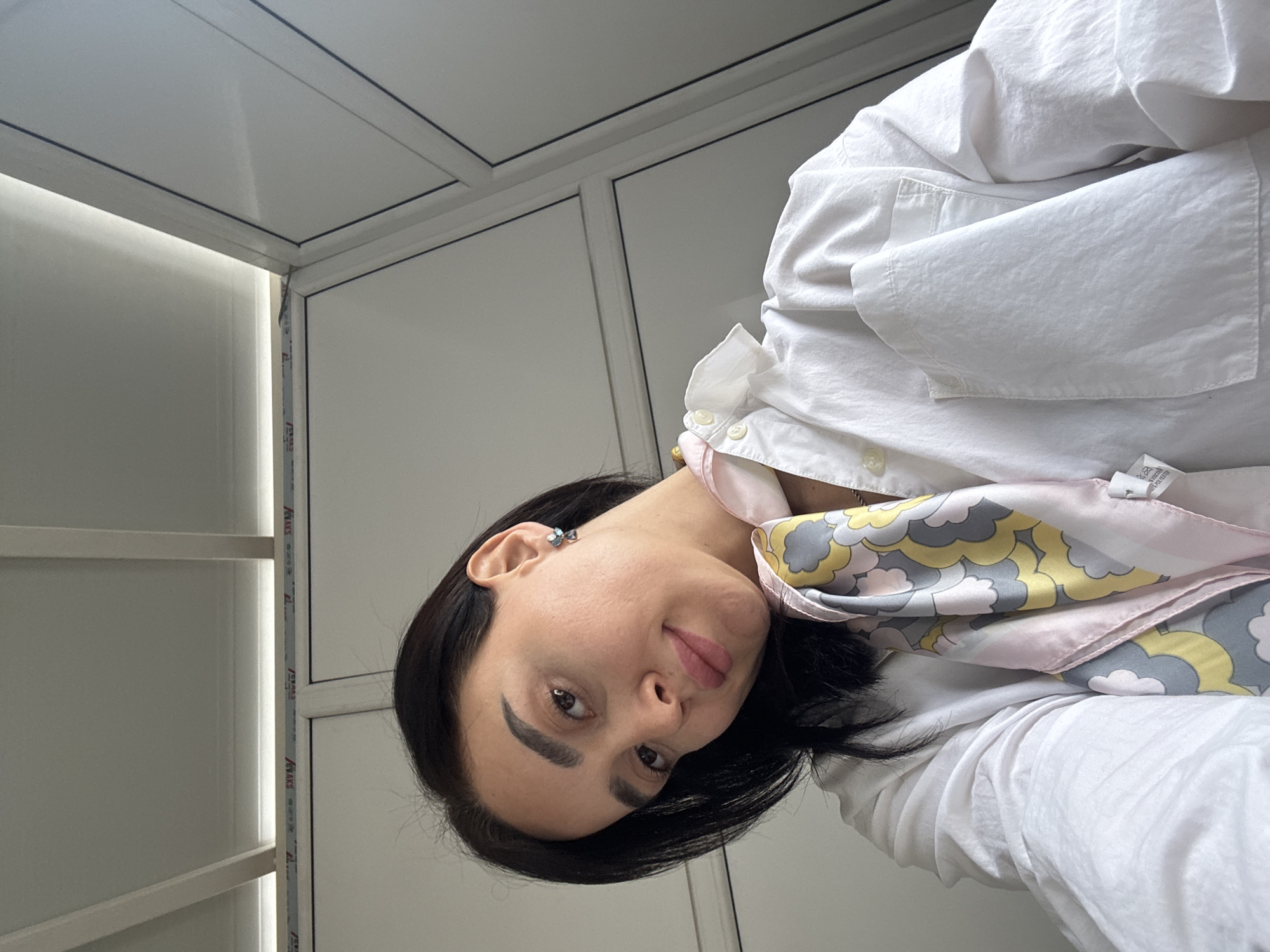 A woman with dark hair is positioned in an interior space. She is wearing a white collared shirt and a patterned scarf. The background features a grid-patterned ceiling and a wall with visible aluminum framing. The image might be taken in a modern office or commercial space. No information about Tbilisi, Georgia is discernible from the visual information provided.