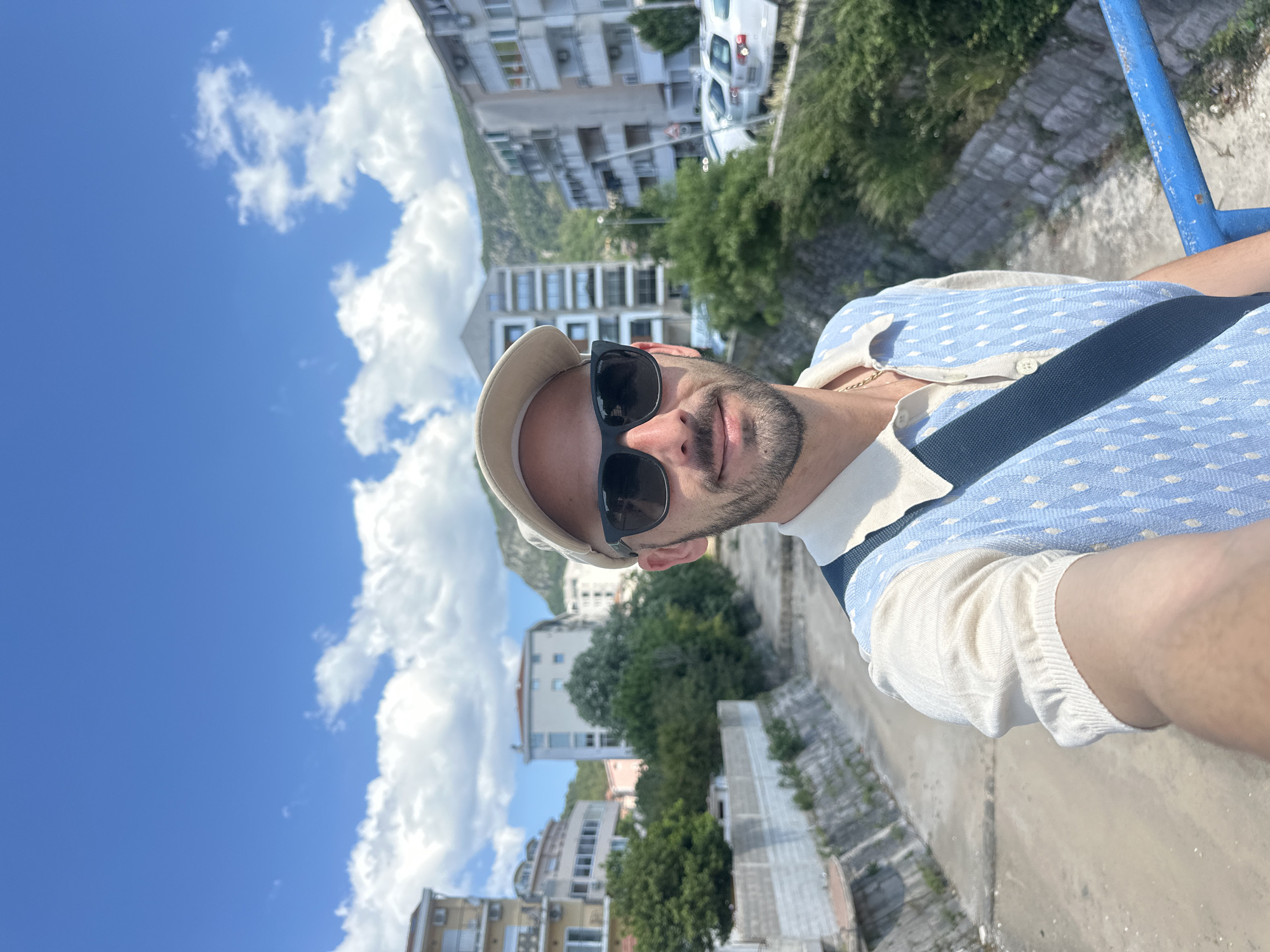 A medium shot depicts a man with a slight beard and sunglasses, wearing a light-colored cap and a blue patterned shirt, seemingly taking a selfie on a bridge in Budva, Montenegro. A dark strap, possibly from a bag, crosses his chest. Buildings and lush greenery fill the background, with a clear blue sky punctuated by fluffy white clouds. A portion of a blue metal railing is visible on the right edge of the frame. The perspective suggests the man is elevated, with the buildings and landscape appearing below.
