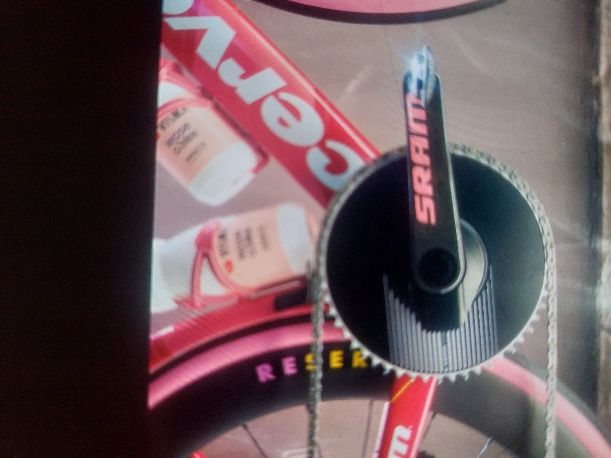 The image features a close-up of a pink bicycle, likely indoors. The bike's frame is labeled "Cervélo" in white letters. Near the frame are two small bottles labeled "Rose Cookie" in white text. The wheel has the word "RESERVE" in mixed pink and yellow letters on the rim. A black SRAM crankset is prominently displayed in the center of the image, with "SRAM" in pink letters on the arm. A chain is visible, connecting the crankset to the rear wheel. The background is blurred. There is no indication that this image was taken in Tsarevo, Bulgaria.