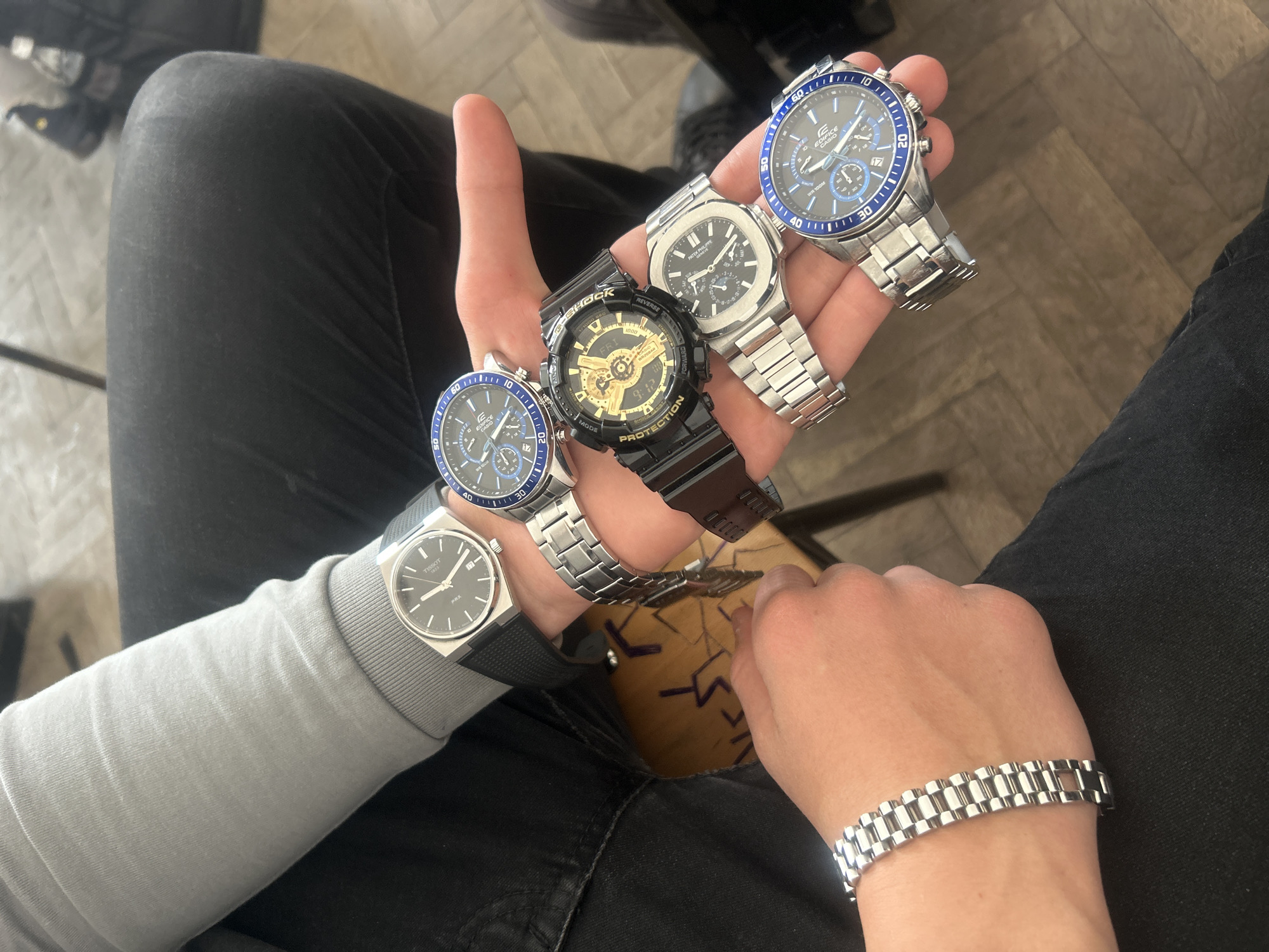The image captures a close-up, eye-level perspective of a person showcasing a collection of watches indoors, likely in Zlatograd, Bulgaria. The individual is wearing black trousers and a light grey long-sleeved top. Their left hand is held open, displaying five different wristwatches, while their right wrist sports a silver-toned metal bracelet resembling a watch band.
The watches displayed on the left hand include:
1. On the wrist, a Tissot PRX with a silver case, black dial, and black rubber strap, displaying "TISSOT PRX".
2. Next to it, a silver Casio Edifice with a blue bezel and black dial on a metal bracelet, with text like "EDIFICE CASIO 100M WR 100M" and numbers "10, 20, 30, 40, 50, 60" visible on the bezel.
3. In the center, a black G-Shock with gold accents, showing "G-SHOCK", "REVERSE", "PROTECTION", "1000", "FORWARD", "MODE", and "FRI 9:12".
4. Further right, a Patek Philippe Nautilus-style watch (possibly a replica) with a silver case and textured dial, showing "PATEK PHILIPPE GENEVE".
5. At the far right, another silver Casio Edifice with a blue bezel and black dial on a metal bracelet, similar to the other Edifice.
The background features a light brown wooden floor with a herringbone pattern. A dark-colored item, possibly a shoe or bag, is partially visible in the top left corner. The activity is clearly the presentation or display of a watch collection. No distinct emotions are visible as the person's face is not shown.
