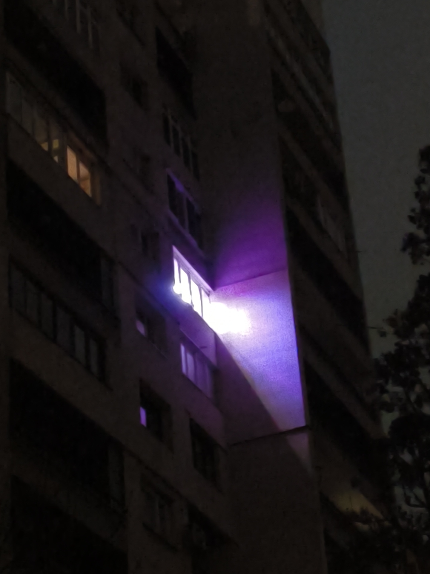 Here is a description of the image:
The image shows a tall building at night. The building has many windows, some of which are lit up. A beam of bright purple light is emanating from one of the windows, shining downwards. To the right of the building, some tree branches are visible against the dark sky. The building appears to be a residential apartment building. I do not know the location in the city of Sofia, Bulgaria.