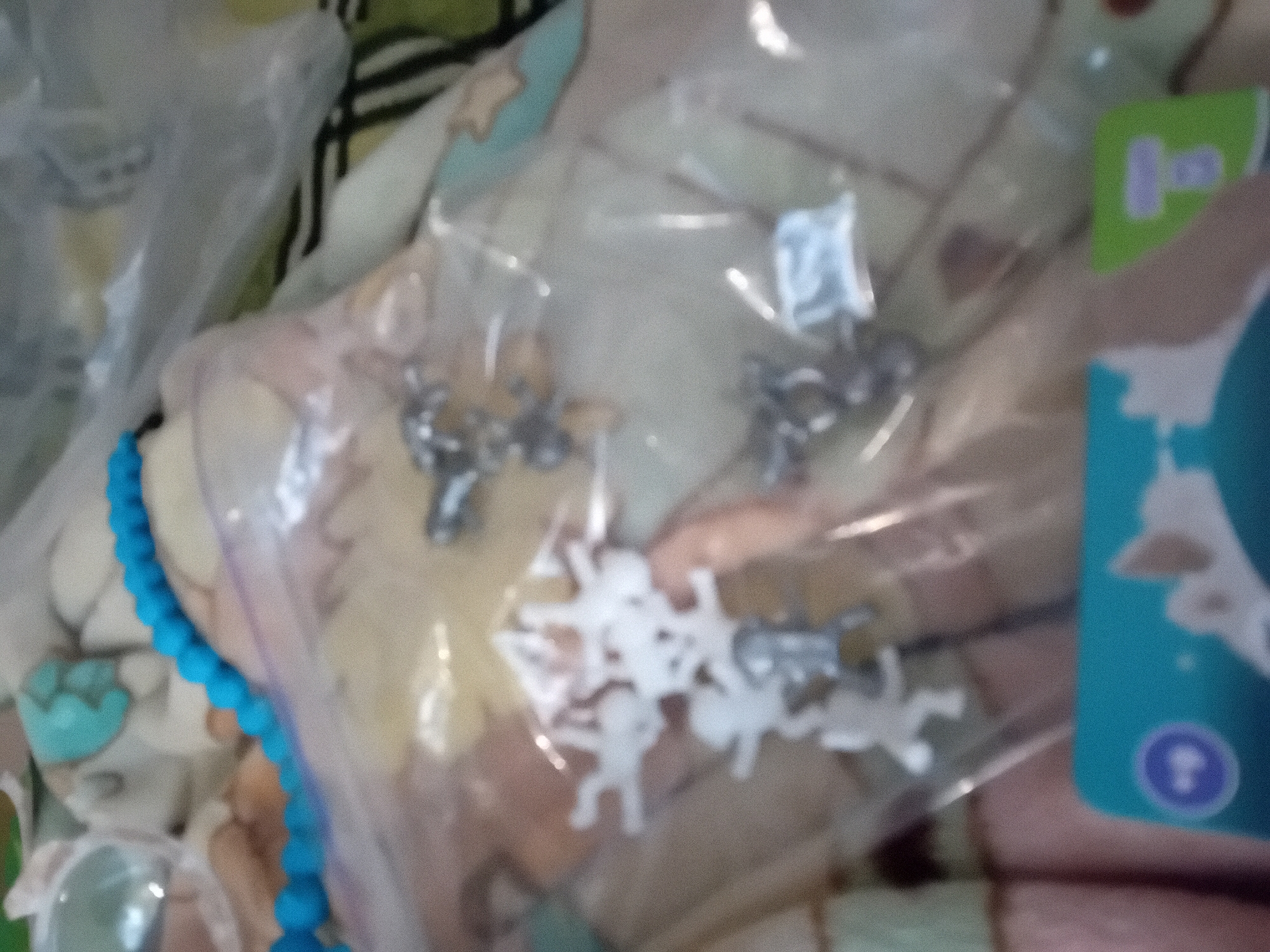 The image shows a close-up of a plastic bag containing several small, white, toy figures. These figures appear to be human-shaped, possibly with outstretched arms and legs. They are scattered within the bag, some overlapping each other. Also visible in the bag are some small, metallic-looking figures, one of which resembles a four-legged animal. A blue beaded string is draped across the top of the bag, and a portion of a blue card with white graphics is visible at the bottom of the image. The background is out of focus, suggesting a soft surface, possibly fabric, with light and dark patterns. There is no discernible information in the image that would allow for the identification of its location in Chuhuiv, Ukraine.