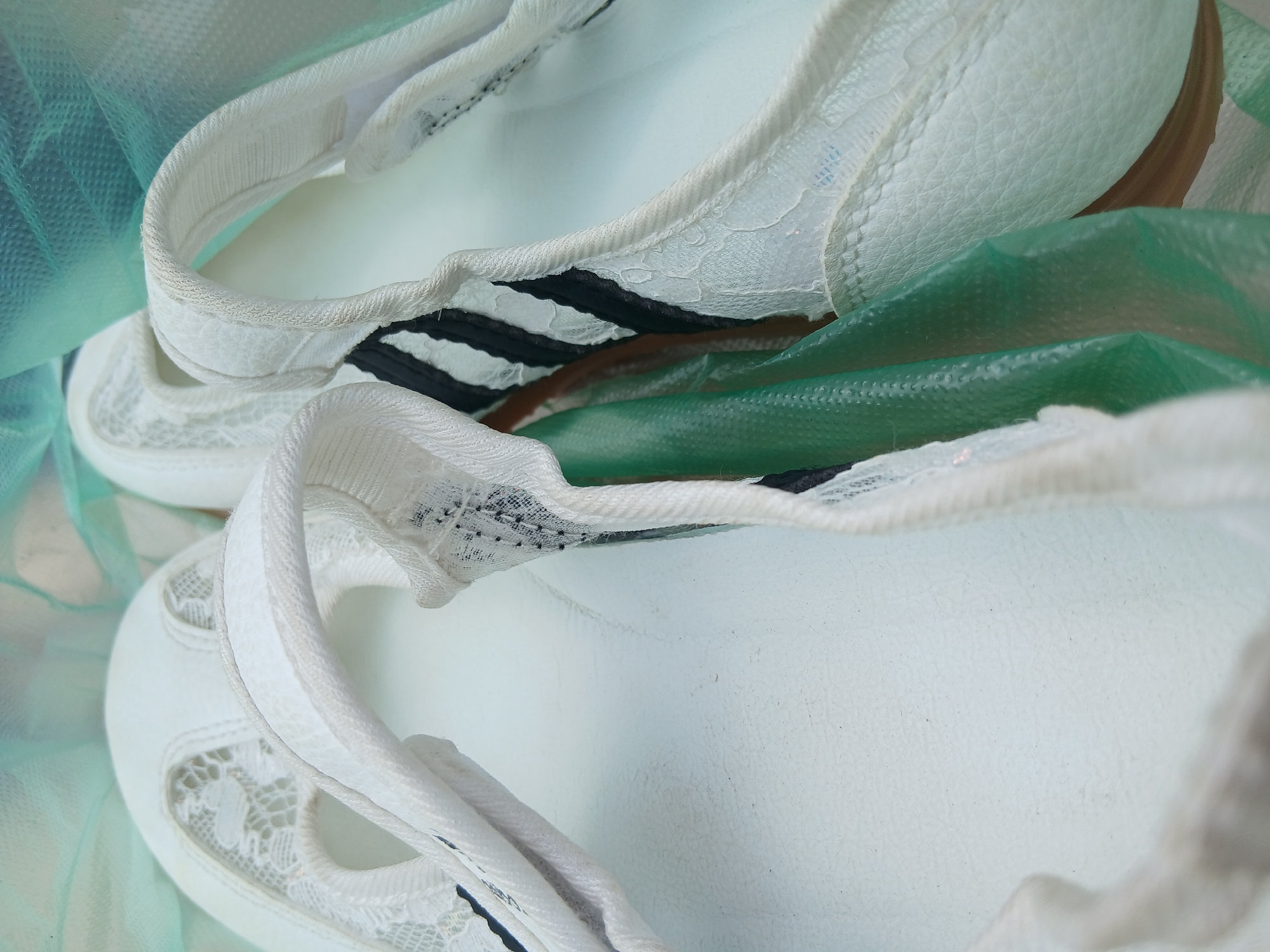The image is a close-up, overhead shot of a pair of white ballet flats. The shoes are made of a textured material, possibly faux leather, and feature white lace inserts on the toe area and along the sides. Black and white striped ribbon accents are visible on the inner side of the shoes. The shoes are placed on a white surface, with a crumpled green plastic material and a sheer, light blue fabric in the background. The lighting is soft and diffused. There are no people or signs of activity in the image. The location is not discernible from the image itself.