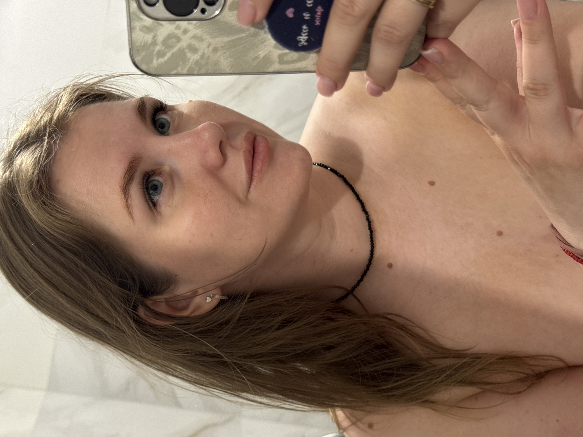 A selfie shows a woman with light brown hair and fair skin. She has blue eyes and appears to be wearing light makeup. She is wearing a black beaded necklace. The photo appears to be taken indoors, with a bathroom-like background of white marble. A smartphone with a gray patterned case and a pop socket is visible as she holds it to take the picture. The specific location within Ферма, Russia, cannot be determined from the image.