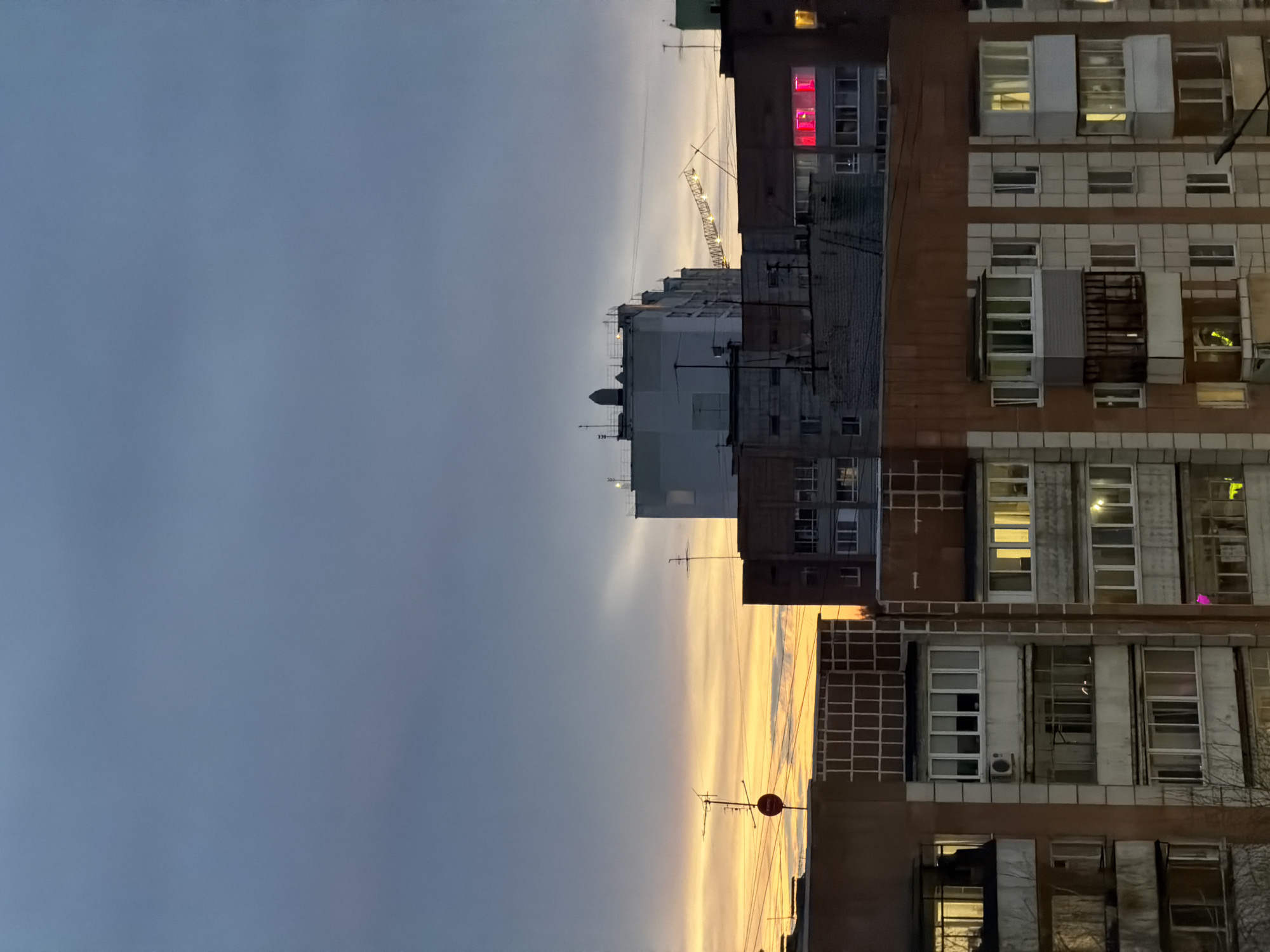 The image depicts an urban scene, likely in the city of Perm, Russia, at dusk or dawn. Buildings are visible, one clad in a brown brick facade and another constructed with grey materials. The building with the brown facade has multiple windows, some with lights on, indicating occupation. A tower crane is located in the foreground; it appears to be under construction. The sky is gradient, dark and cloudy above, with a band of golden light near the horizon. Antennas are present on the building and atop adjacent buildings. The scene suggests an urban environment with active construction during the transition of day and night.