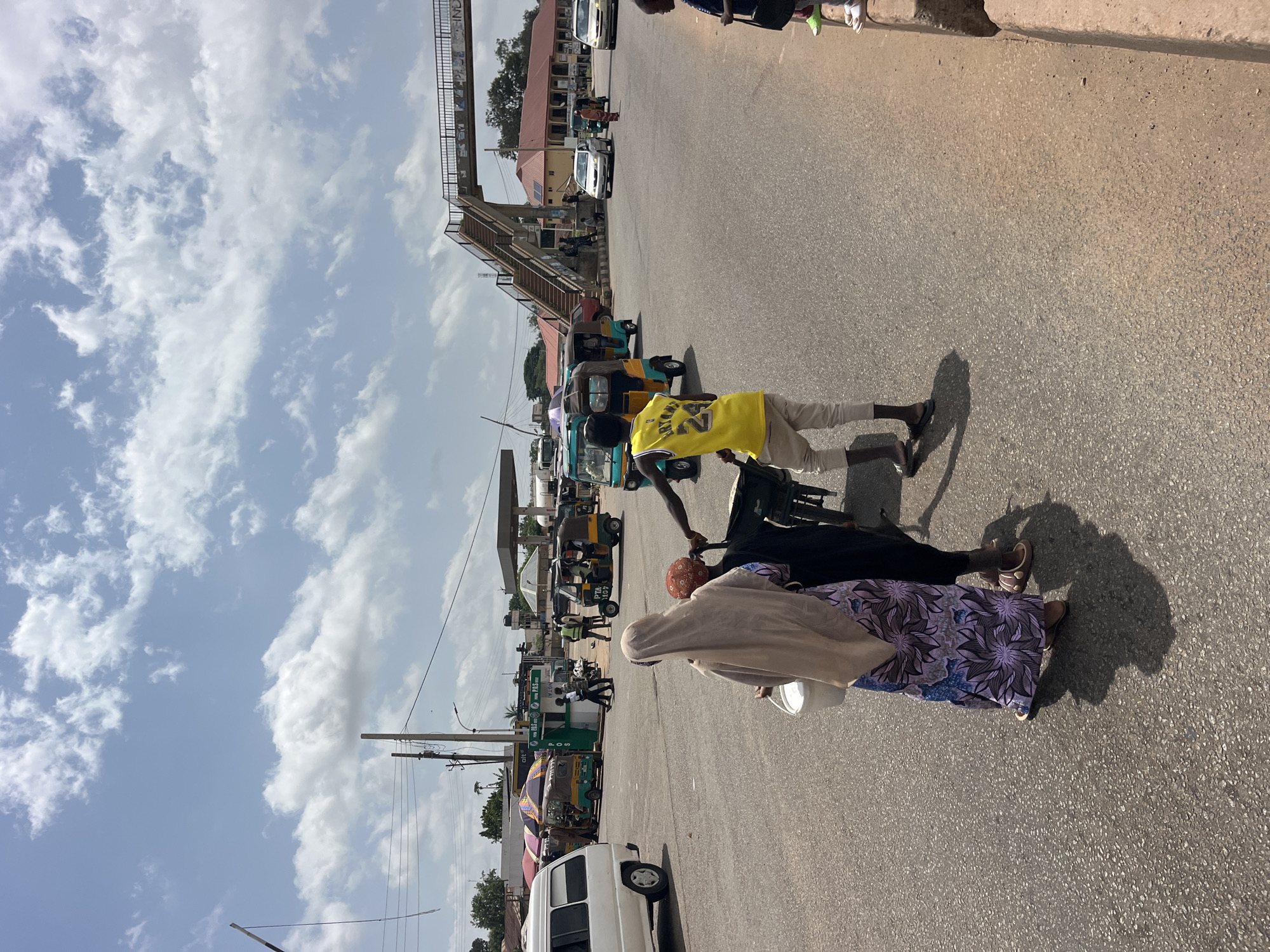 The image shows a street scene in a location that appears to be a town or city in a sunny climate. The orientation of the image is vertical, with the sky taking up the upper portion and the ground the lower portion.
In the foreground, three individuals are visible. One person, wearing a yellow t-shirt with a basketball player's number on it, is pushing a cart or trolley. Another person, dressed in a light-colored headscarf and a patterned dress, is walking alongside the cart. A third person, mostly obscured, is also near the cart. Their clothing suggests warm weather.
The background reveals a street with several vehicles, including what appear to be auto-rickshaws or tuk-tuks, and a white van. There is a pedestrian overpass in the distance, indicating a busy intersection or road. Buildings line the street, some with reddish roofs, and there are signs visible on some of the structures, though the text is not legible. Power lines and poles are also present.
The sky is partly cloudy, with patches of bright blue visible between white cumulus clouds. The sunlight is strong, casting shadows on the ground, suggesting it is daytime, likely midday or afternoon. The overall impression is of a lively, bustling street environment. There is no clear indication of the specific location or any discernible emotions from the people depicted.