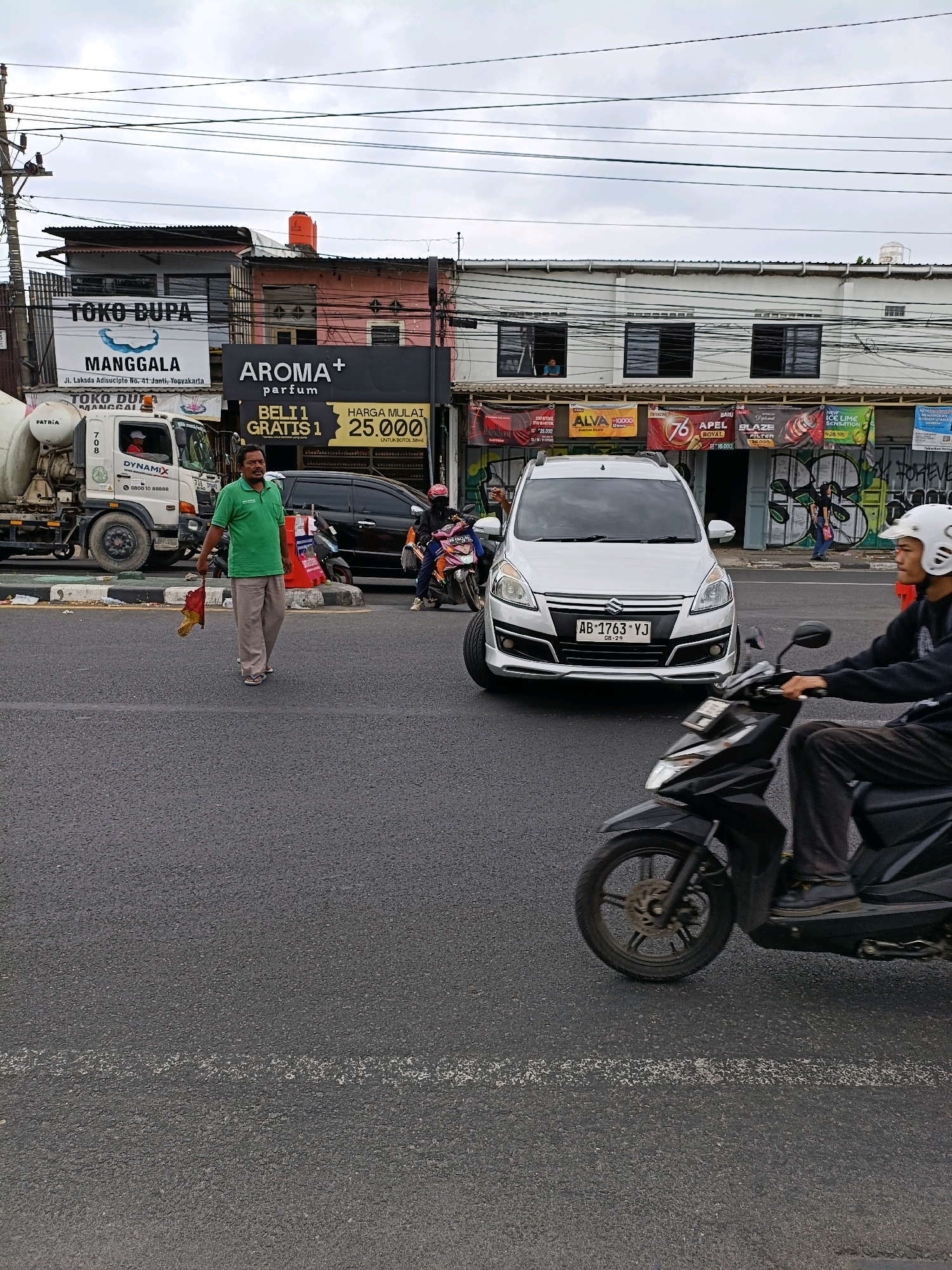 A man in a green shirt and khaki pants, holding a red and yellow flag, walks across an asphalt road in front of a white SUV with an Indonesian license plate AB 1763 YJ. He appears to be directing traffic or signaling. A black motorcycle with a rider wearing a white helmet is in the foreground, to the right of the SUV. In the background, buildings with storefronts line the street. One storefront displays a banner for "AROMA+ parfum" with a promotion for "BELI 1 GRATIS 1" and a price of 25,000. Another banner advertises "TOKO DUPA MANGGALA." A cement mixer truck is parked to the left of the man. The sky is overcast, and there are power lines overhead. The location is identified as Depok, Indonesia, based on context.