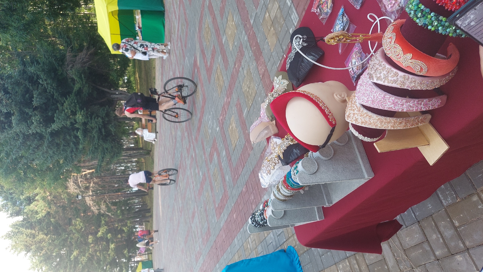 The image depicts an outdoor market scene, likely a fair or festival, set against a backdrop of lush green trees. In the foreground, a table is laden with various accessories, including a mannequin head wearing a red headband adorned with gold trim, several other headbands embellished with intricate beadwork in shades of pink, orange, and gold, and racks displaying an assortment of beaded bracelets in vibrant colors like green and red. The table is covered with a deep red cloth.
In the mid-ground, several individuals are visible. Two people are riding bicycles on a paved walkway. One cyclist, wearing a white t-shirt and dark shorts, rides an orange bicycle. Another cyclist, wearing a red backpack, rides a black bicycle. Further back, a person in a white outfit sits on a bench. Adjacent to them, two individuals are standing near a yellow and green tent, possibly a vendor's stall, where goods are displayed on tables.
The environment suggests a public park or promenade, with the ground paved with interlocking bricks in a red and gray diamond pattern. The overall atmosphere appears to be casual and recreational, with people enjoying a sunny day outdoors. The presence of a market suggests a local event in Komsomolsk-on-Amur, Russia, likely held during warmer months given the attire of the people and the lush greenery.