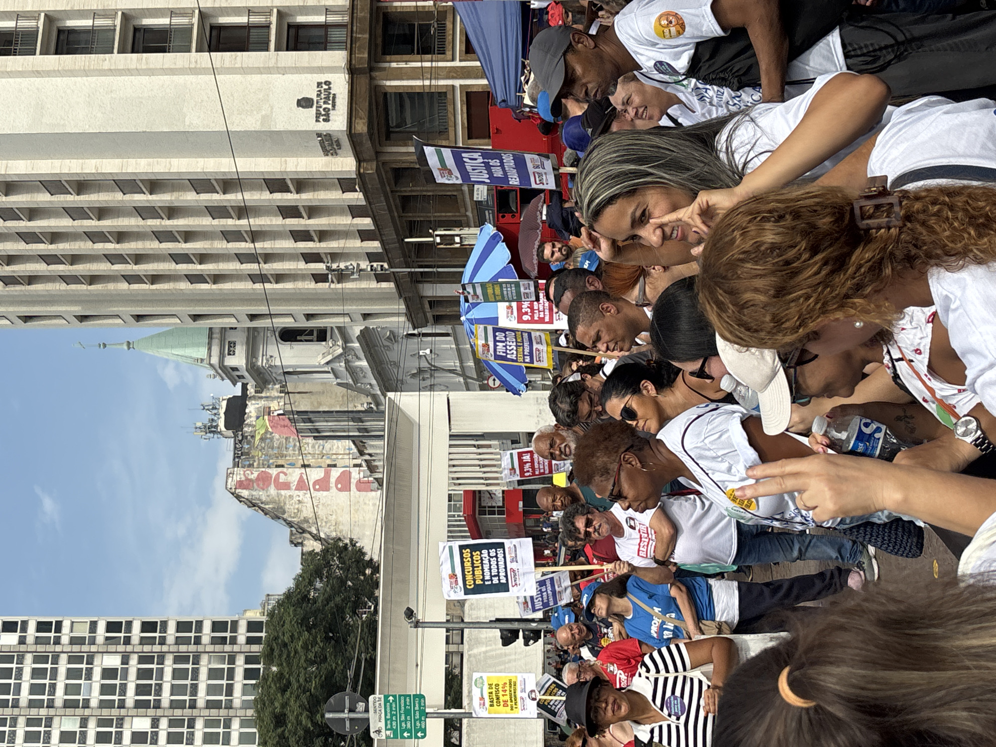 The image captures a political rally in the city of São Paulo, Brazil. A large crowd of people is gathered in a plaza or street, with several holding signs expressing various demands. Some signs mention "Concursos Públicos" (Public Competitions) and "Nomeação de Todos os Aprovados" (Appointment of All Approved), suggesting a focus on public sector employment. Another sign says "Fim do Assédio Sexual e Moral na Prefeitura" (End Sexual and Moral Harassment in the City Hall). Other signs include "Justiça Para os Readaptados" (Justice for the Readapted). The crowd appears to be diverse, with people of different ages and ethnicities present.
The backdrop includes the architecture of São Paulo, with a building bearing the inscription "Prefeitura de São Paulo" visible. Also, a mural is visible on a building. The sky is partly cloudy, indicating daylight conditions. The general atmosphere appears to be one of protest and demonstration, with people advocating for specific causes.