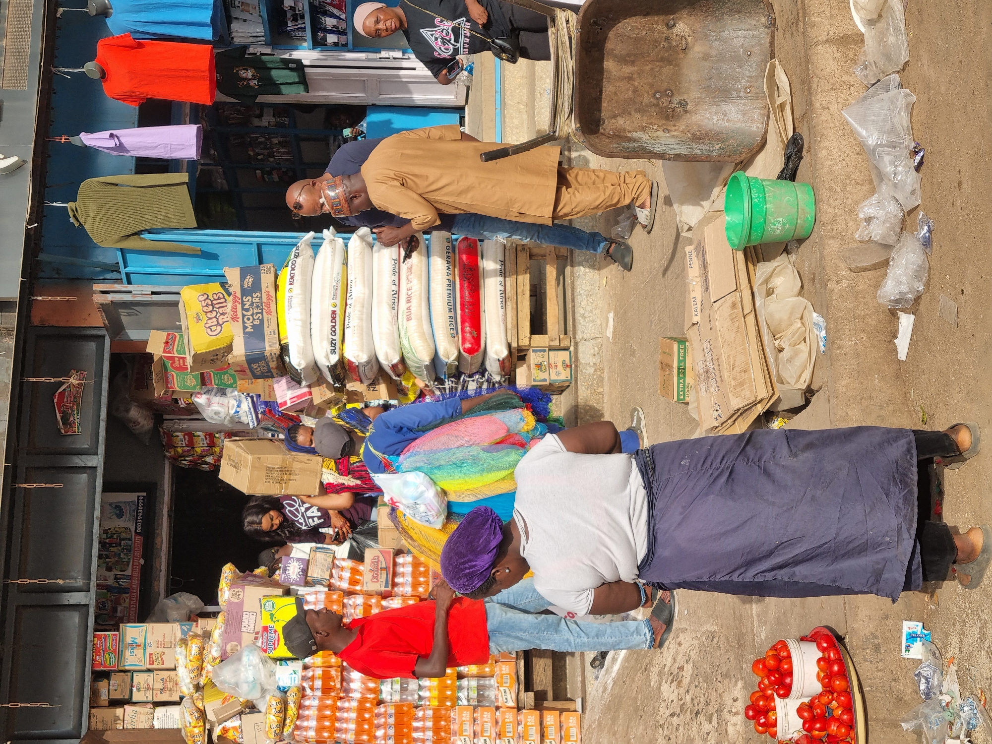 The image depicts a bustling street scene outside a shop at Wokwo Plaza, Ahmadu Bello Way, in Jos, Nigeria. The shop's front is packed with goods, including stacks of packaged food items, drinks like Maltina, and bags of rice. Several people are present. One man in a red shirt is positioned amidst stacks of drinks, while another, wearing a brown traditional outfit and hat, stands near the bags of rice. Two women are seen nearby. Clothing items hang above the shop, adding color. A wheelbarrow and a green bucket rest on the pavement, surrounded by plastic bags and boxes.