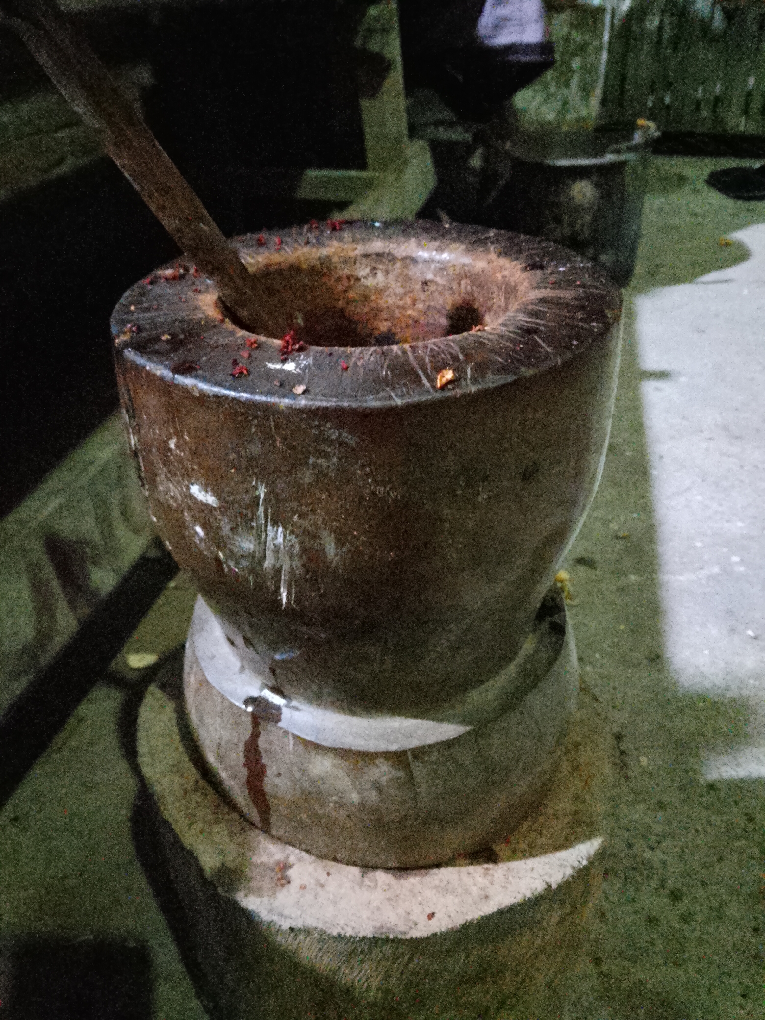 A weathered, stone mortar with a wooden pestle is positioned on a circular wooden base. The mortar is dark brown and appears to be made from a coarse-grained material, with visible scratches and white markings on its exterior. Small red specks are scattered around the rim and inside the mortar, suggesting it has been used for grinding spices or other ingredients. The wooden pestle, dark and worn, is angled into the mortar. The mortar and its base sit on a rough, textured surface, possibly concrete or stone, with patches of dark green and lighter grey. Shadows cast long, dark lines across the ground. In the background, out of focus, are other dark, cylindrical objects and what appears to be a dark wall or structure. The lighting is dim, suggesting it is nighttime or an indoor space with minimal illumination. There is no indication of the specific location within the city of Payakumbuh, Indonesia.