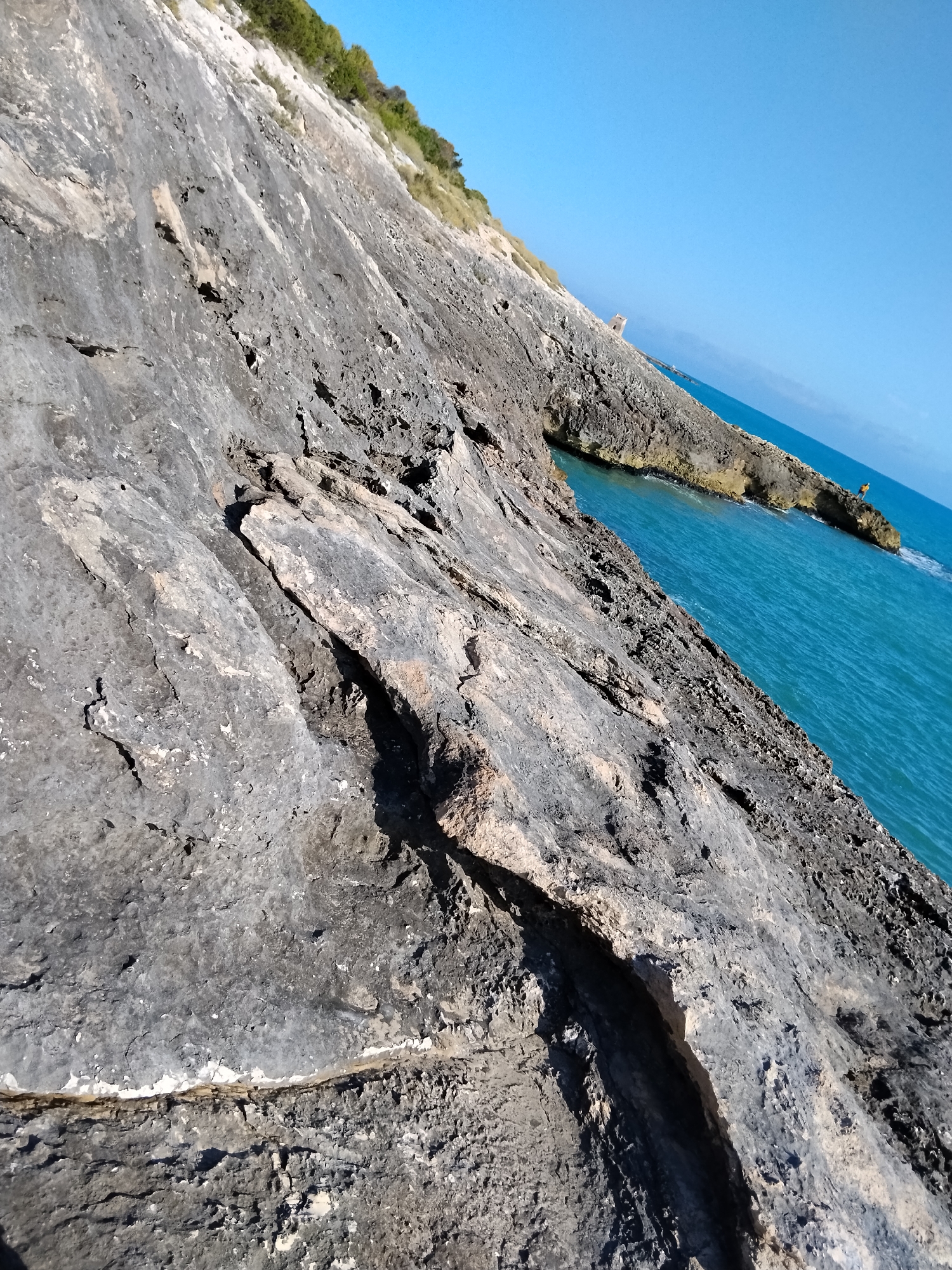 The image displays a rugged, rocky coastline under a clear blue sky. The foreground is dominated by a steep, grey rock face with visible fissures and textures, angled diagonally from the bottom left to the top right. Beyond the rocks, the bright blue sea stretches to the horizon. In the middle ground, a small rocky outcrop juts into the water, with a single person standing on it. Further out, a hazy landmass can be seen on the horizon. The overall scene suggests a natural, coastal environment. This is likely the coastline of Vieste, Italy, known for its dramatic sea stacks and rocky shores.