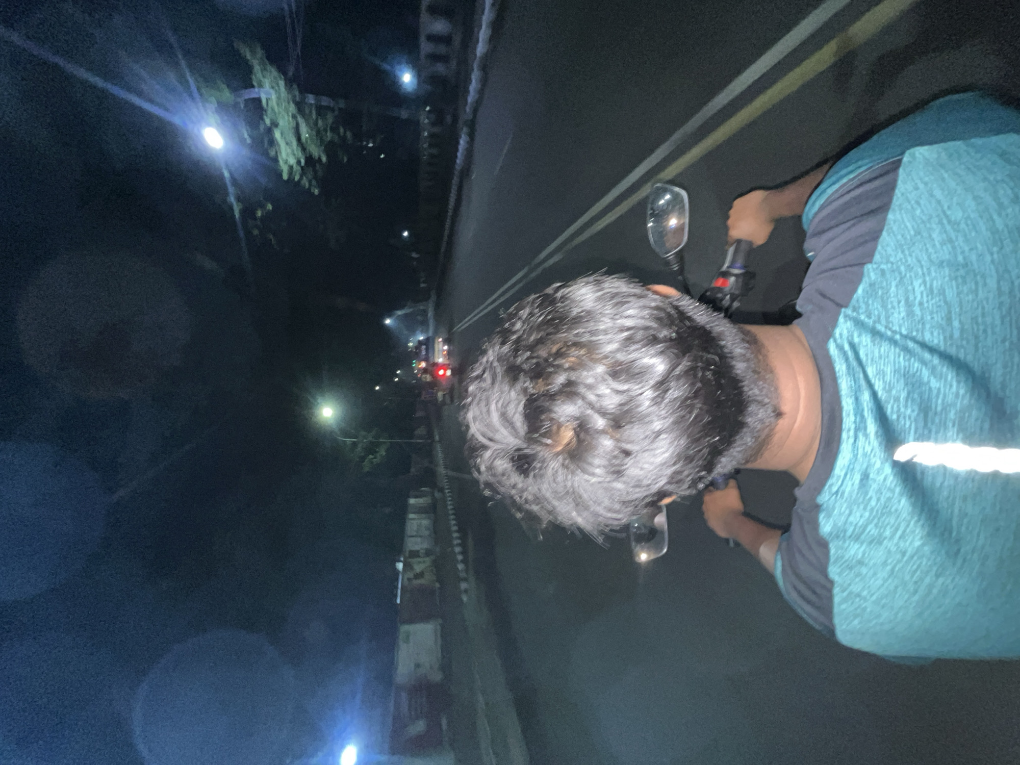 The image shows a person riding a motorcycle at night. The scene appears to be along a road in the city of Dhaka, Bangladesh, based on the presence of streetlights, buildings, and other vehicles. The person is wearing a blue and grey shirt and has dark hair. The road has lane markings, and the motorcycle's side mirror is visible. The environment is dark, with light sources from streetlights and vehicle headlights. The motorcycle appears to be in motion.