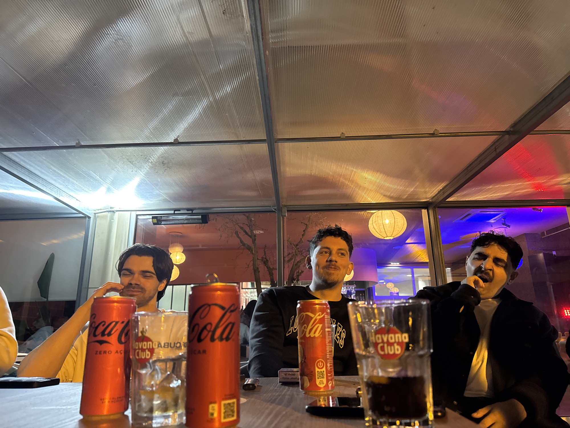 Three young men are seated at a table. On the table are Coca-Cola cans and glasses with the words "Havana Club". The location is Vila Real, Portugal. The scene appears to be inside a covered patio or enclosed outdoor dining area with clear roofing and glass walls. The area is illuminated with hanging lights and outside the enclosure, there are trees and urban structures with lit signage. One man is touching his face. Another man appears to be yawning or talking. The third man is looking straight ahead.