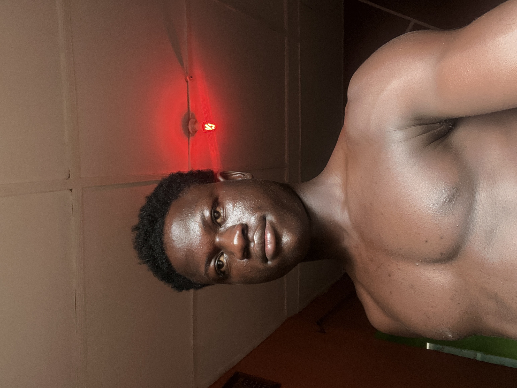 A dark-skinned man is seen in an indoor setting. He is shirtless, exposing his upper torso and chest. The lighting appears to be dim, with a red light visible on the wall. The walls are painted in light shades, possibly beige or white. The specific location within the city of Mosafejaw, Nigeria, cannot be determined from the visual information provided.
