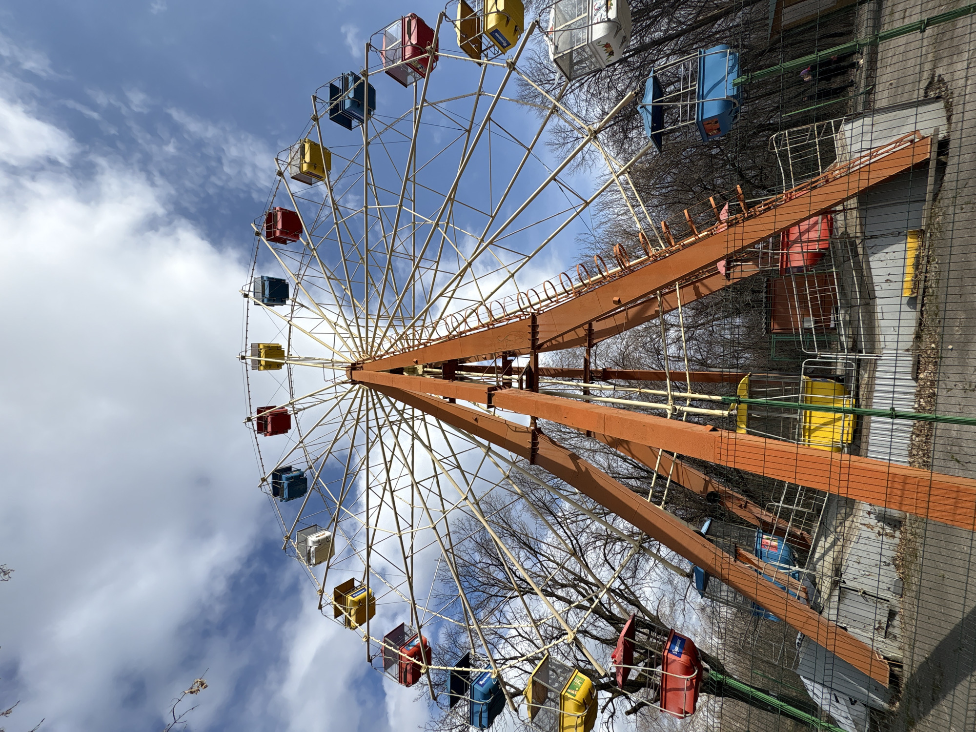 The image depicts a Ferris wheel in Ulyanovsk, Russia. The wheel is made of orange-painted metal with passenger cars in various colors: red, yellow, blue, and white. The sky is blue with scattered white clouds. Bare trees are visible behind the wheel, suggesting it may be late autumn or early spring. The wheel is not in motion. A chain-link fence is partially visible at the right side of the image.