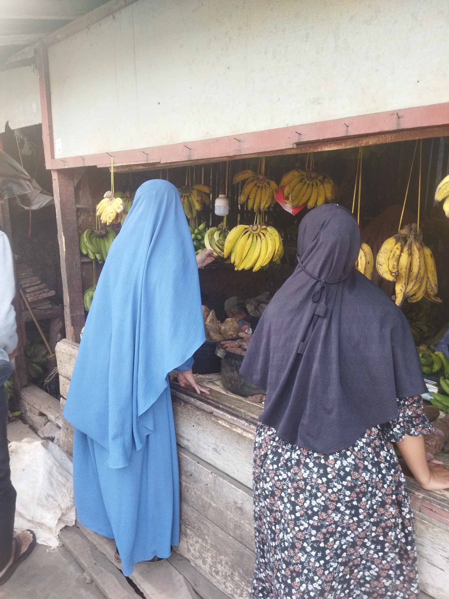 Two women wearing hijabs are standing at a fruit stand in Banjar, Indonesia. The woman on the left is wearing a long blue hijab and dress, and the woman on the right is wearing a dark hijab and a floral print dress. They are both looking at the bananas displayed at the stand. The fruit stand appears to be outdoors, with wooden planks forming the structure. Bunches of ripe bananas hang from hooks above the women. The ground is made of dirt and some wooden planks. The overall scene suggests a local market or roadside stall.