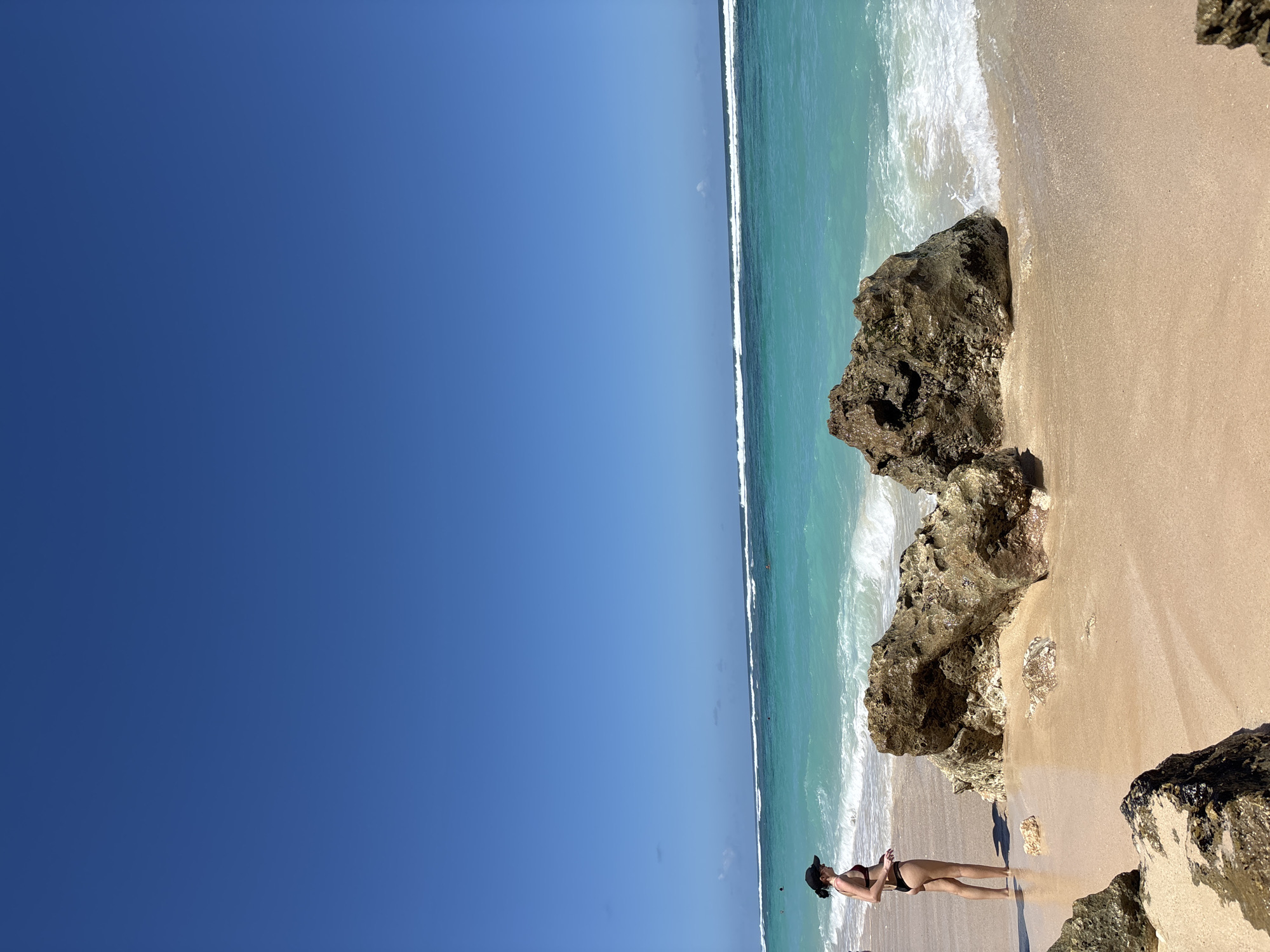 The image depicts a coastal scene in Kuta Selatan, Indonesia. A woman in a dark-colored bikini stands on a sandy beach near rocky formations. The turquoise ocean, characterized by white-crested waves, meets the shoreline. A clear, blue sky occupies a large portion of the frame. The lighting suggests a sunny day.