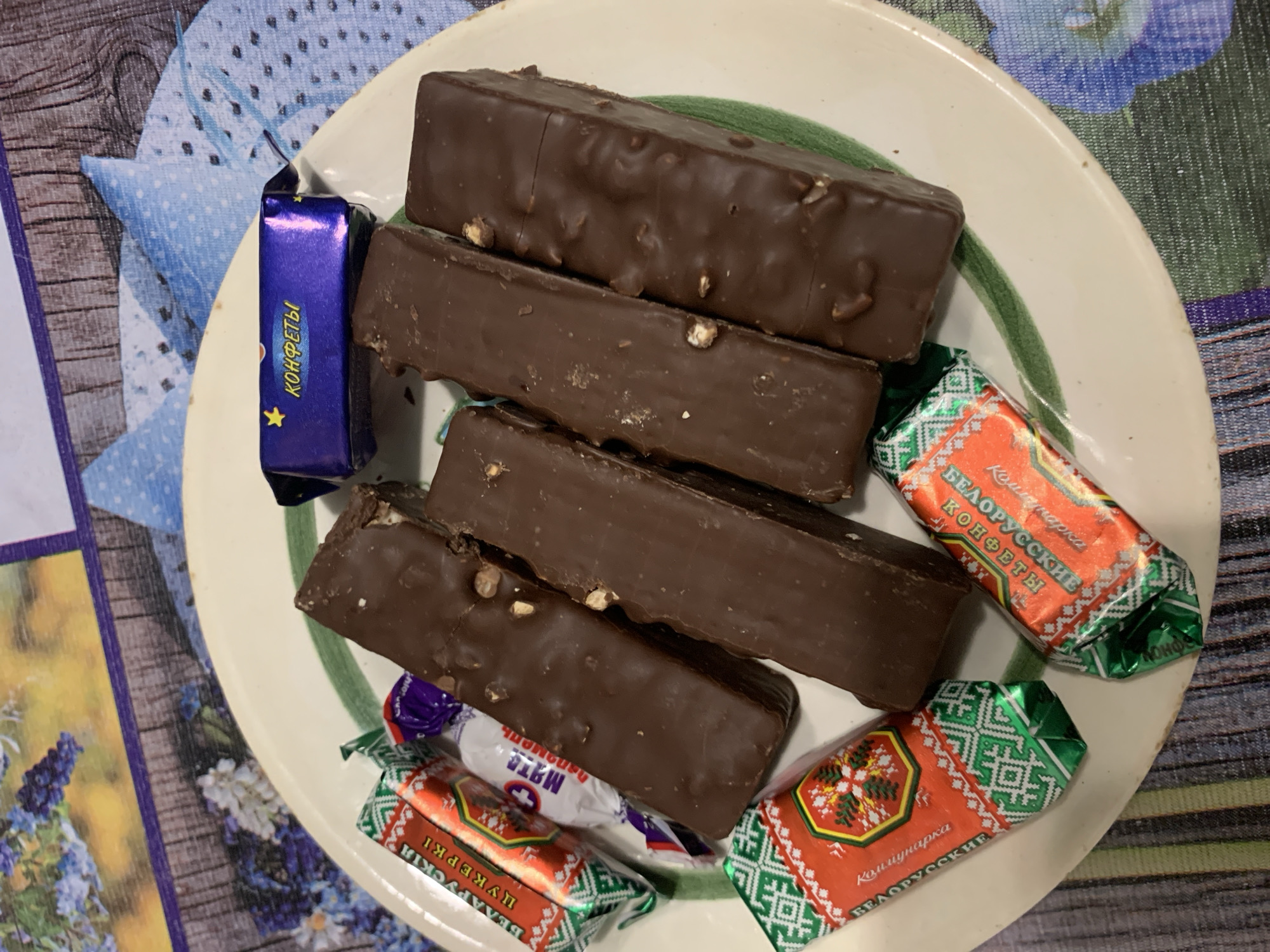 This is a top-down, close-up shot of a plate of assorted chocolates. The plate is white with a subtle green rim. There are six chocolate bars, all covered in dark chocolate and some appear to have small nut pieces embedded in them. Several individually wrapped candies are also visible. One candy wrapper is blue and says "КОНФЕТЫ" (Confectionery) in yellow Cyrillic lettering. Two other candies are wrapped in red and green foil featuring a snowflake-like pattern and Cyrillic text that appears to be in Belarusian. The chocolates are arranged on a patterned tablecloth, which has elements of blue, purple, and green. The background shows a blurred floral and wooden pattern. The scene is likely indoors, possibly in a kitchen or dining area. There is no indication of location in Відзаўскі сельскі Савет, Belarus.